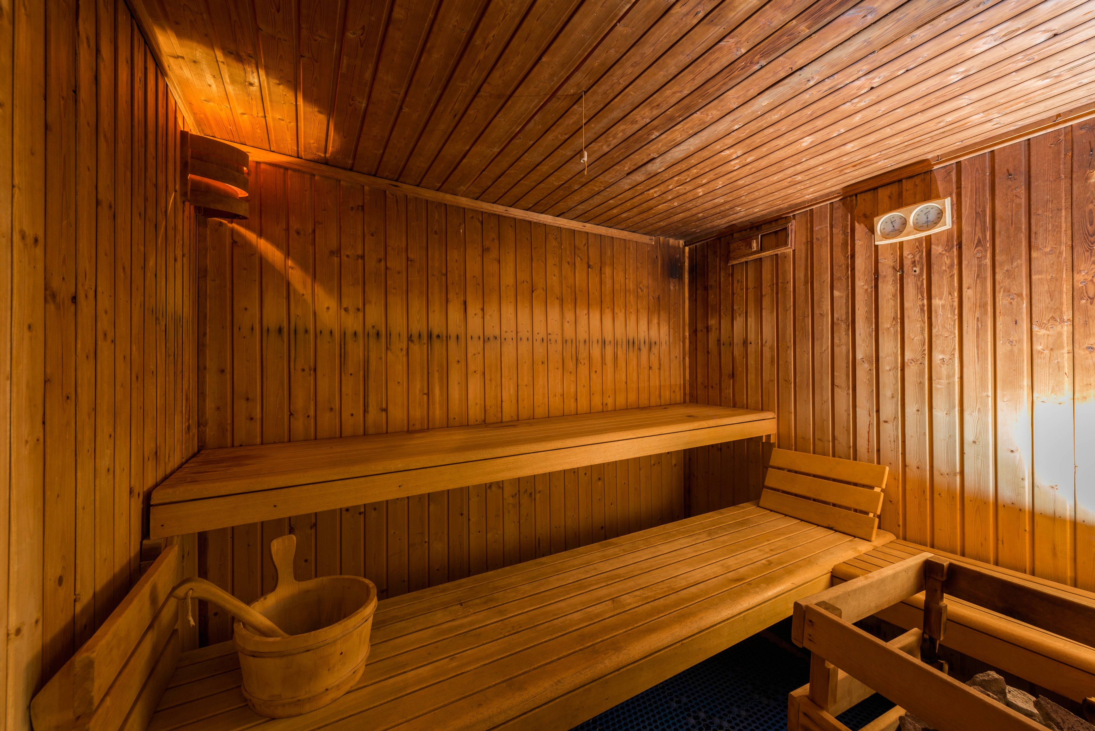 Warm wooden sauna interior featuring tiered benches and soft lighting. A wooden bucket with a ladle rests on the lower bench, emphasizing traditional sauna accessories. The walls and ceiling are lined with vertical wood panels, creating a cozy, rustic atmosphere. No people are present, highlighting the calm, relaxing environment.