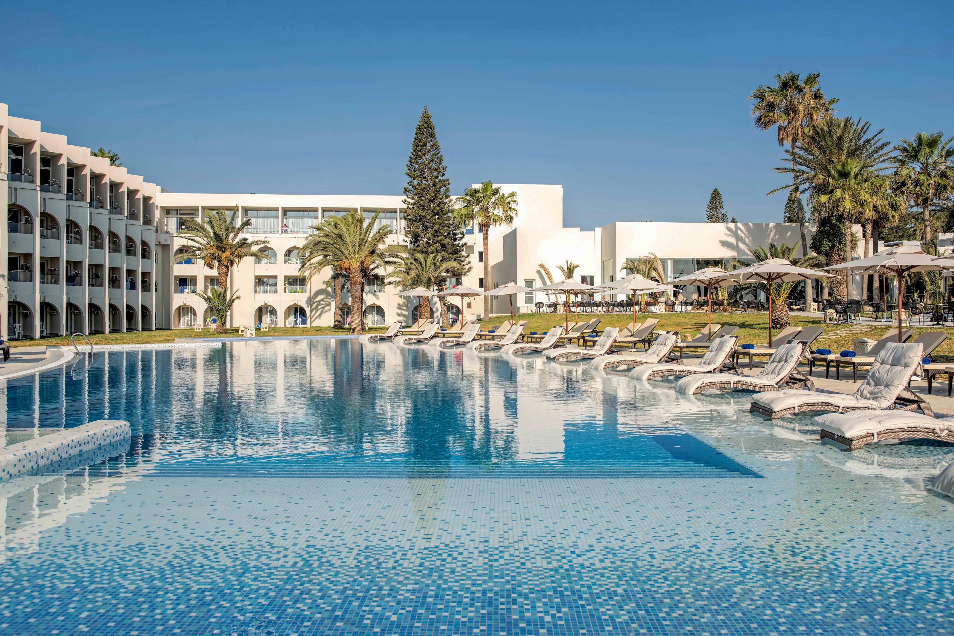 A large outdoor swimming pool stretches across the foreground of a modern resort. White sun loungers partially submerged in the shallow water line the pool edge, facing manicured lawns and palm trees. Curved hotel buildings with balconies frame the left side, while additional white structures and umbrellas appear on the right. The clear blue sky and calm water create a serene, sunny vacation atmosphere.