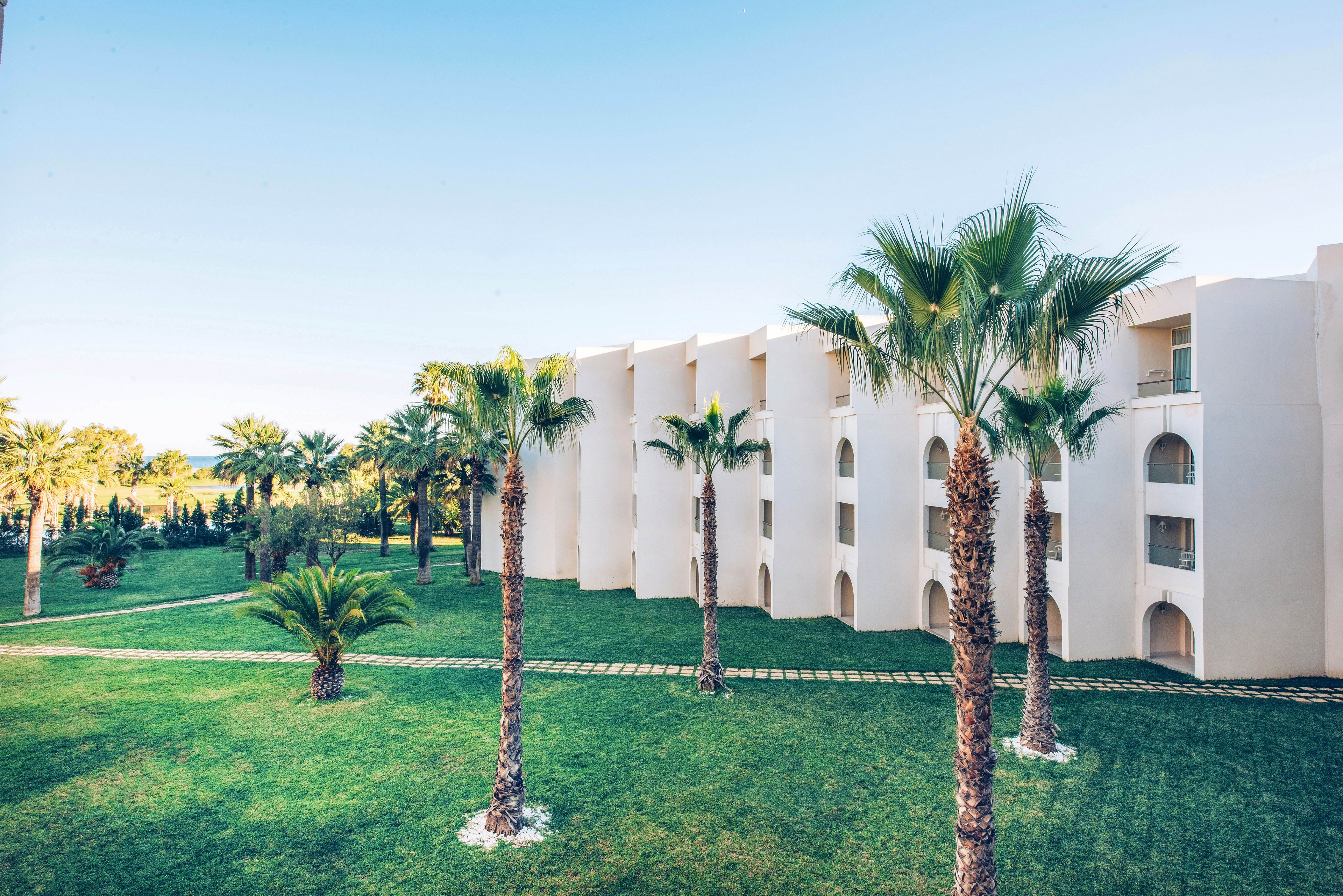 A modern white resort building stands beside a neatly manicured lawn lined with tall palm trees. The architecture features multiple floors with arched balconies facing the green courtyard. Bright daylight and a clear blue sky create a warm, inviting atmosphere. The scene suggests a tranquil vacation destination in a sunny, coastal or tropical region.