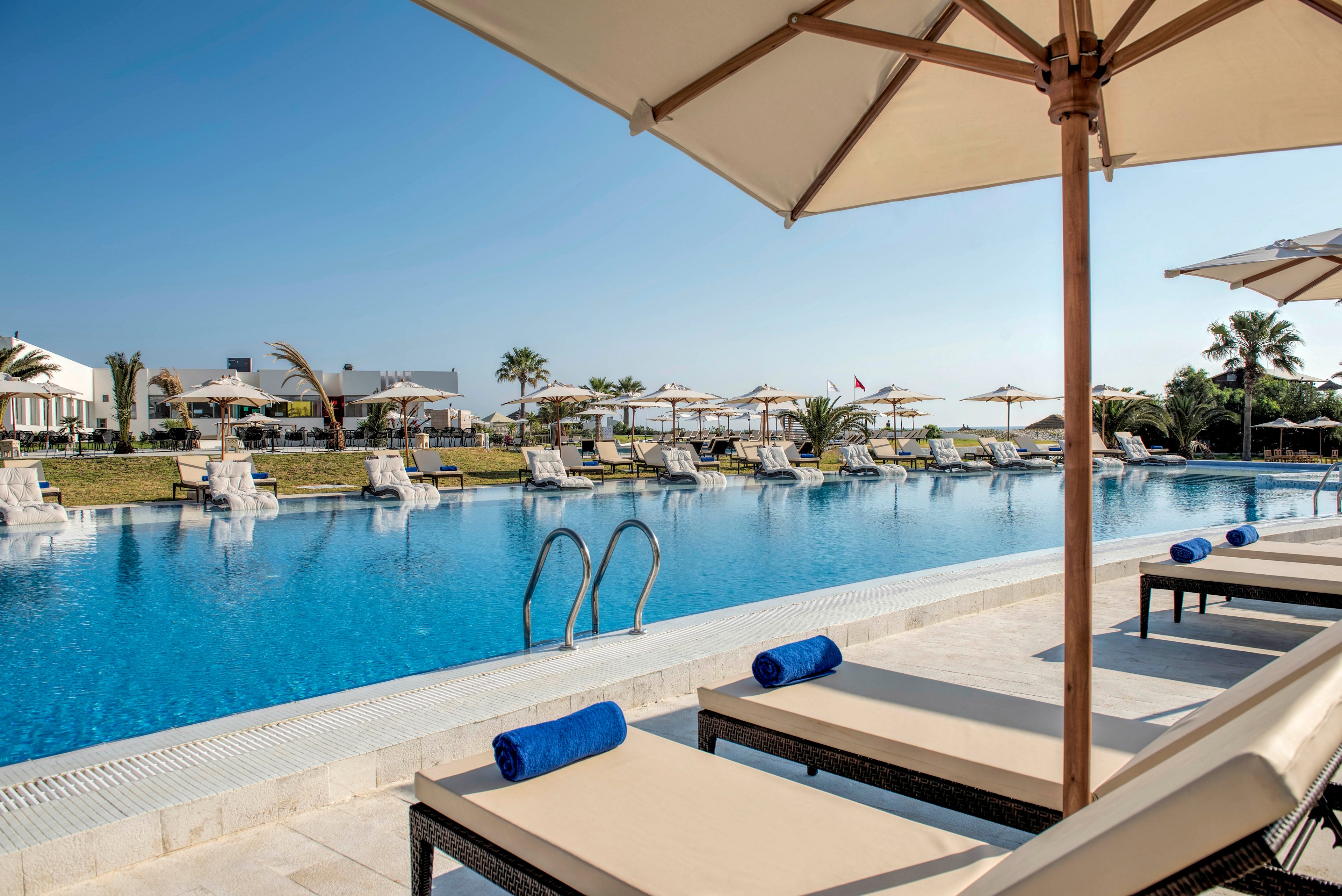 A large outdoor swimming pool stretches alongside a row of cushioned sun loungers at a luxury resort. Beige umbrellas provide shade while blue towels are neatly rolled on the loungers. In the background, additional loungers and umbrellas line the opposite side of the pool, with palm trees and low resort buildings visible. The clear blue sky and calm water create a serene, relaxing vacation atmosphere.