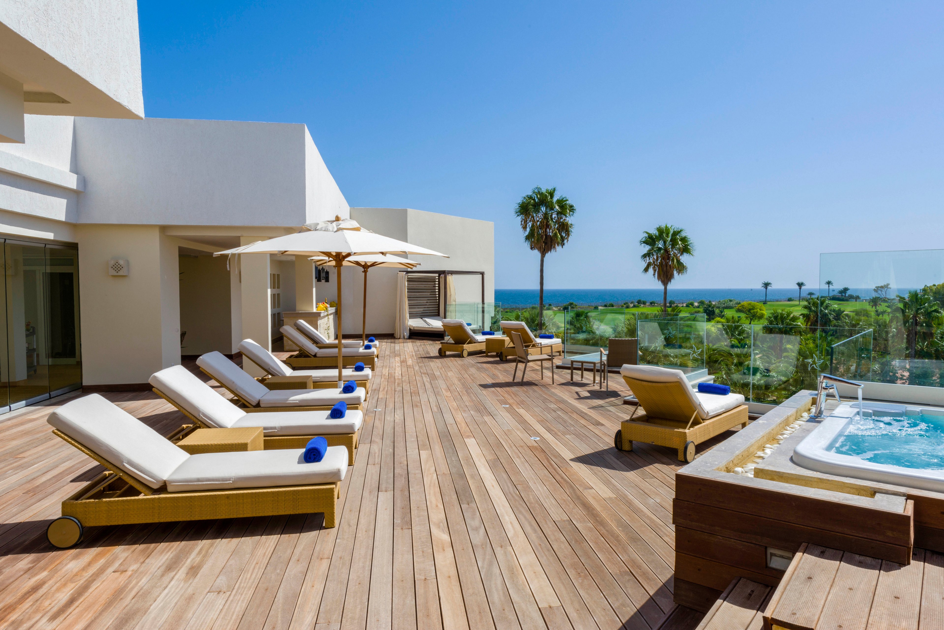 Spacious wooden deck terrace of a modern seaside hotel features multiple sun loungers neatly arranged under large white umbrellas. A bubbling outdoor hot tub sits on the right side, overlooking lush greenery and palm trees. Glass railings frame the terrace, opening to a clear view of the blue ocean and cloudless sky. The scene suggests a calm, sunny day ideal for relaxation and vacation leisure.