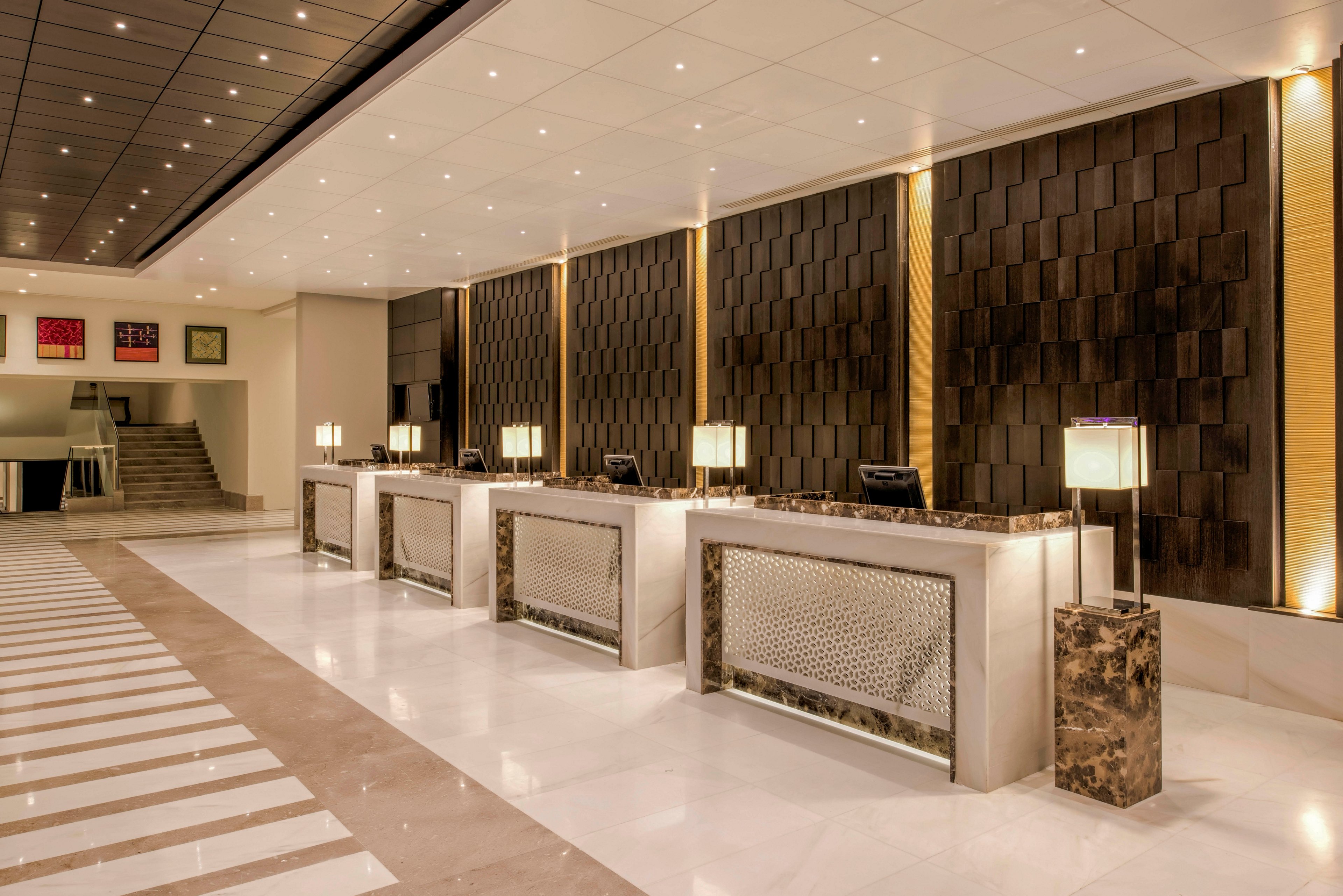 Spacious hotel reception area featuring multiple check-in counters with illuminated front panels. The lobby has a polished marble floor with striped detailing leading toward the desks. Warm lighting, dark textured wall panels, and contemporary table lamps create an upscale, welcoming atmosphere. A staircase and framed artwork are visible in the background, enhancing the modern design aesthetic.