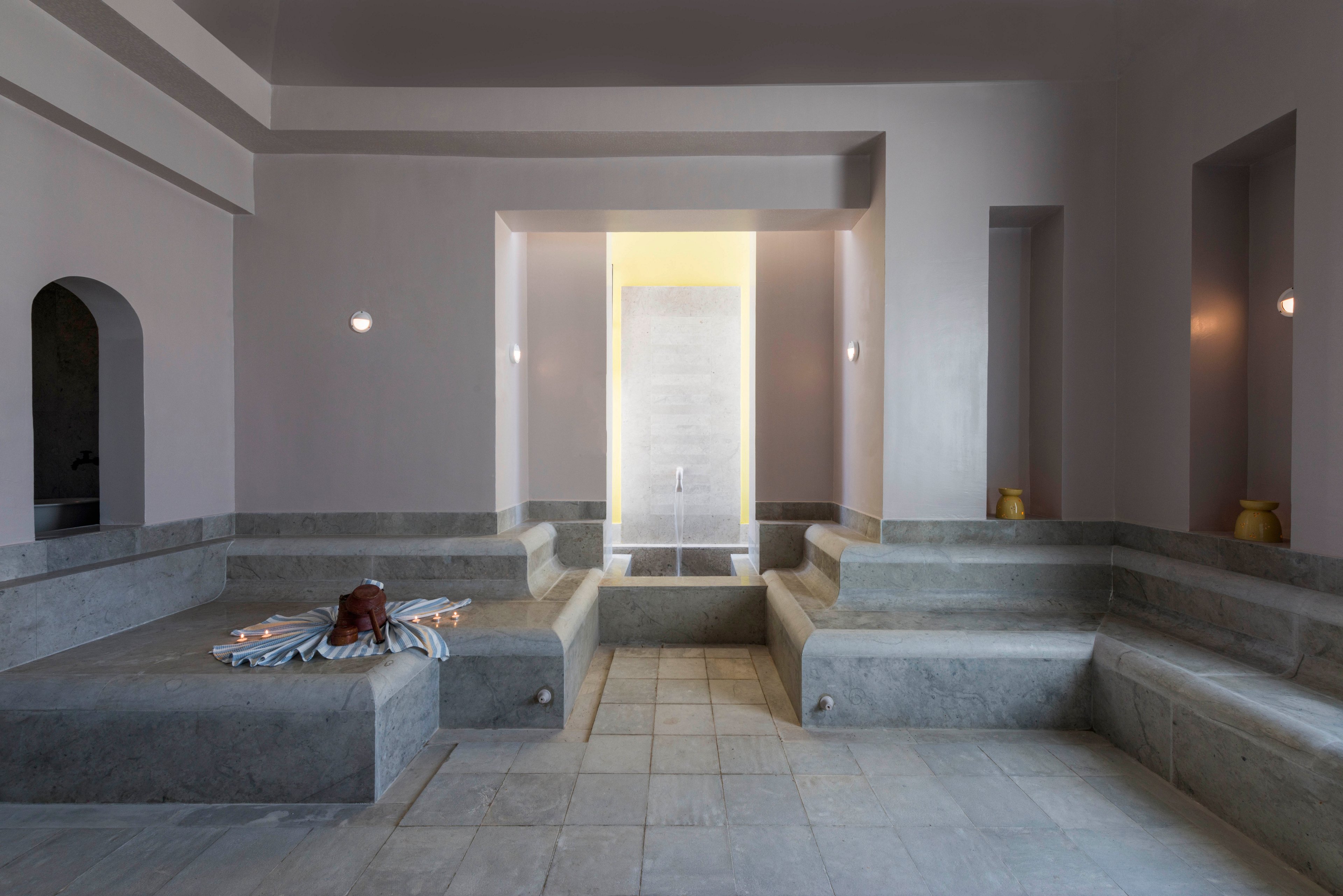 A serene spa hammam interior features wide stone benches and a central recessed water area. Soft, indirect lighting highlights the smooth concrete surfaces and pale neutral walls. A folded towel and bath accessories rest on one of the benches, suggesting a prepared relaxation space. Arched doorways and simple wall sconces contribute to the calm, minimalist atmosphere.