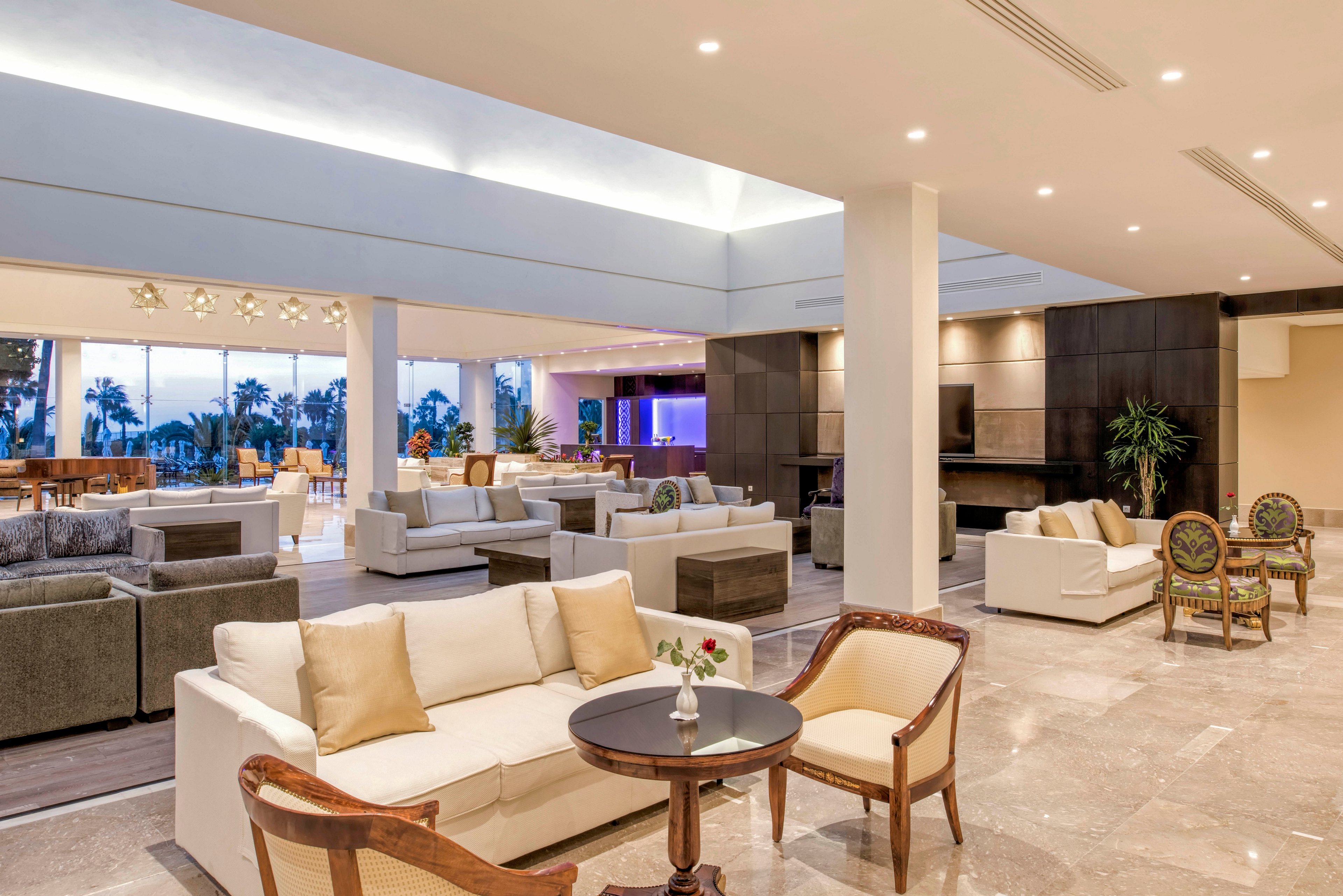 A bright, contemporary hotel lobby features multiple seating areas with sofas and armchairs arranged for conversation. Large windows at the back reveal palm trees and an outdoor view, bringing natural light into the space. Neutral tones of beige, white, and gray dominate the interior, accented by warm wood and subtle lighting. The polished floor and clean architectural lines create an upscale, welcoming atmosphere.