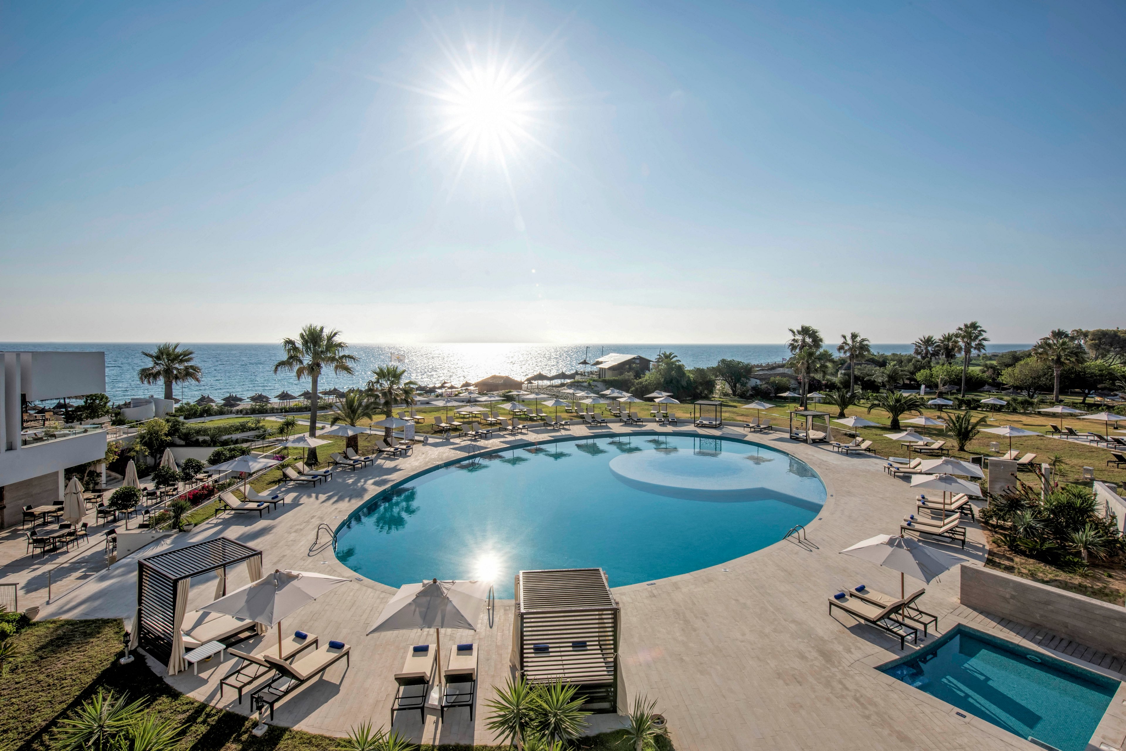 A large circular swimming pool sits at the center of a modern seaside resort. Sun loungers and cabanas are arranged neatly around the pool on spacious terraces. The bright sun shines high in a clear blue sky, casting reflections on the calm water. In the background, landscaped gardens lead down to the coastline and open sea, creating a tranquil vacation atmosphere.