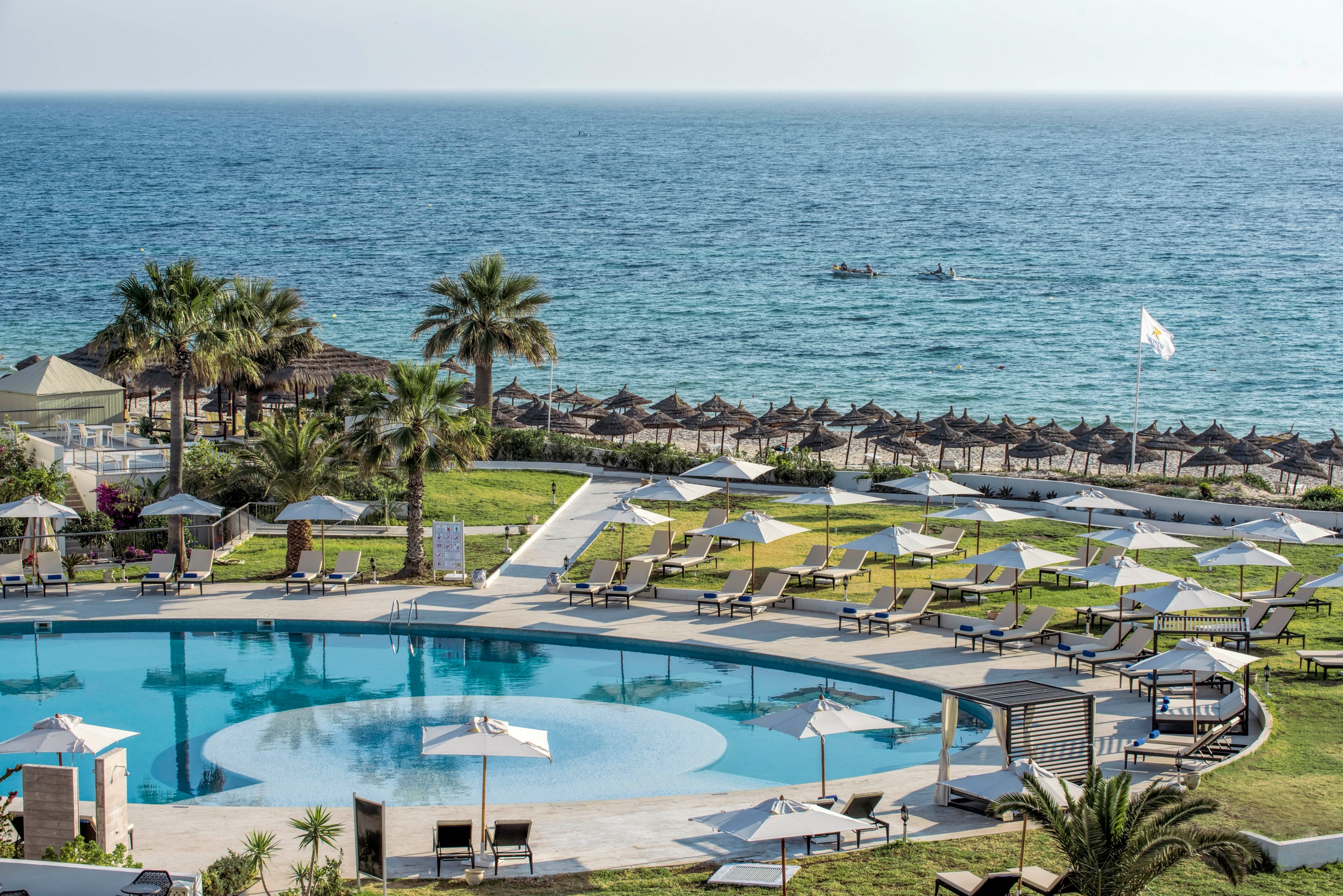 A circular swimming pool with sun loungers and white umbrellas sits in the foreground of a seaside resort. Beyond the pool, manicured lawns and palm trees lead down to a sandy beach lined with rows of thatched parasols. The calm blue sea stretches to the horizon, where a small boat can be seen on the water. The scene suggests a relaxing vacation atmosphere under clear, bright daylight.