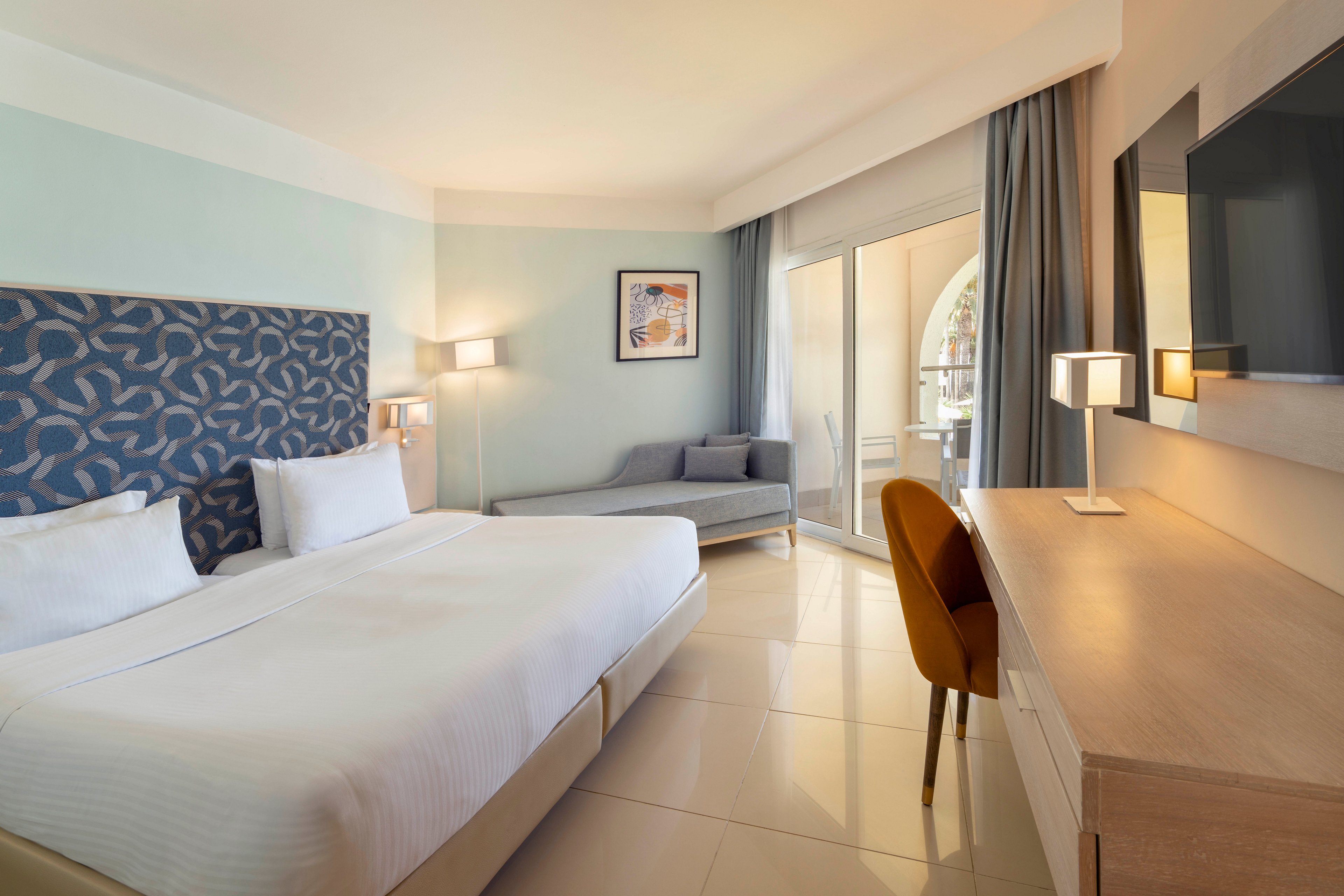 Spacious contemporary hotel room featuring a large double bed with patterned headboard. A sleek wooden desk with chair and table lamps faces a wall-mounted television. The room includes a gray sofa near framed artwork and large sliding glass doors leading to a balcony with arched openings. Soft neutral tones and ample natural light create a calm, inviting atmosphere.