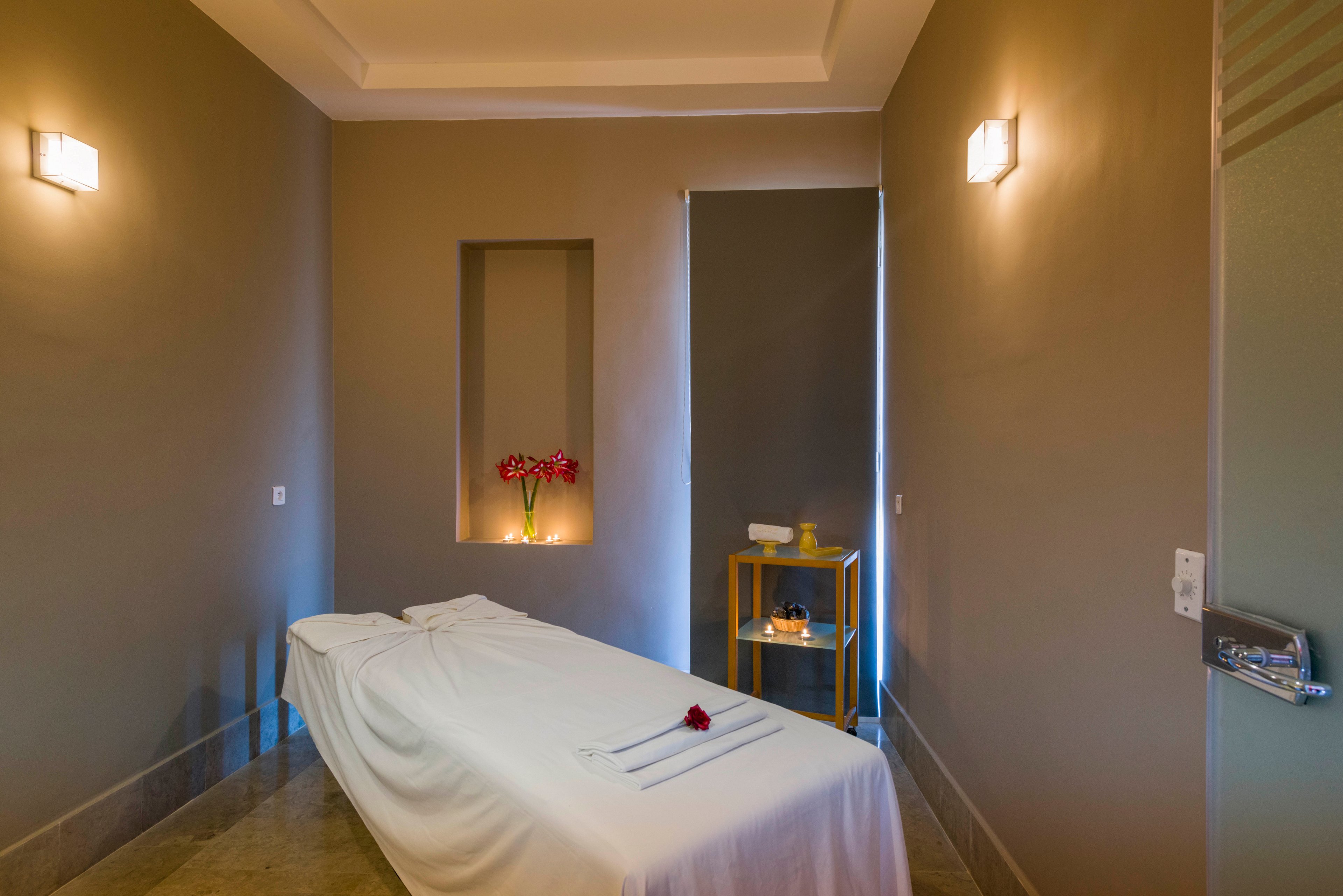 A neatly made massage table covered in white linens stands in the center of a tranquil spa room. Warm wall sconces cast soft light on neutral-colored walls, creating a calm and inviting atmosphere. A recessed niche with flowers and a small side table holding candles and towels add to the relaxing setting. The overall scene suggests a peaceful space for wellness treatments and relaxation.