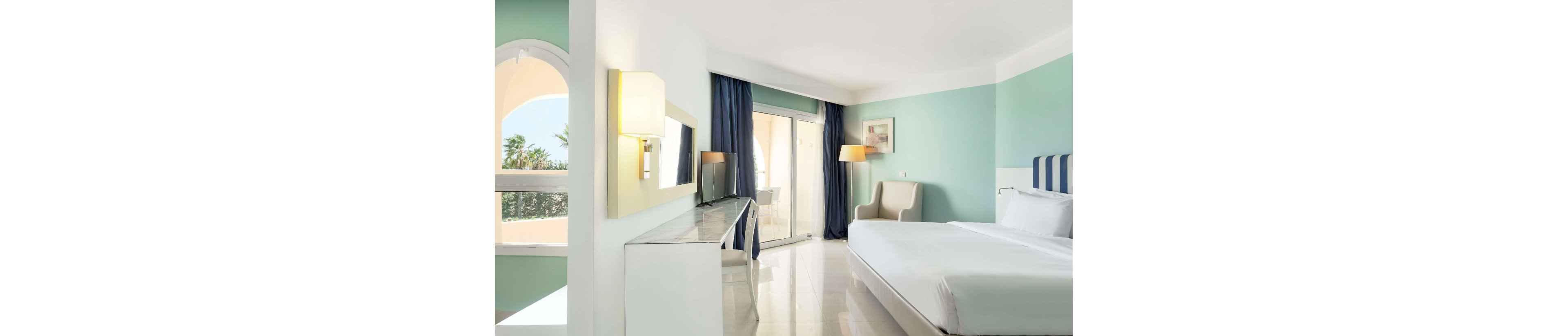 A modern hotel bedroom features a large neatly made bed with a striped headboard against a mint-green wall. The room opens onto a balcony through glass doors framed by dark blue curtains, allowing natural light to flood the space. A sleek desk with a mirror, a contemporary armchair, and warm wall and floor lamps add comfort and style. Soft pastel colors and clean lines create a calm, airy atmosphere ideal for relaxation.