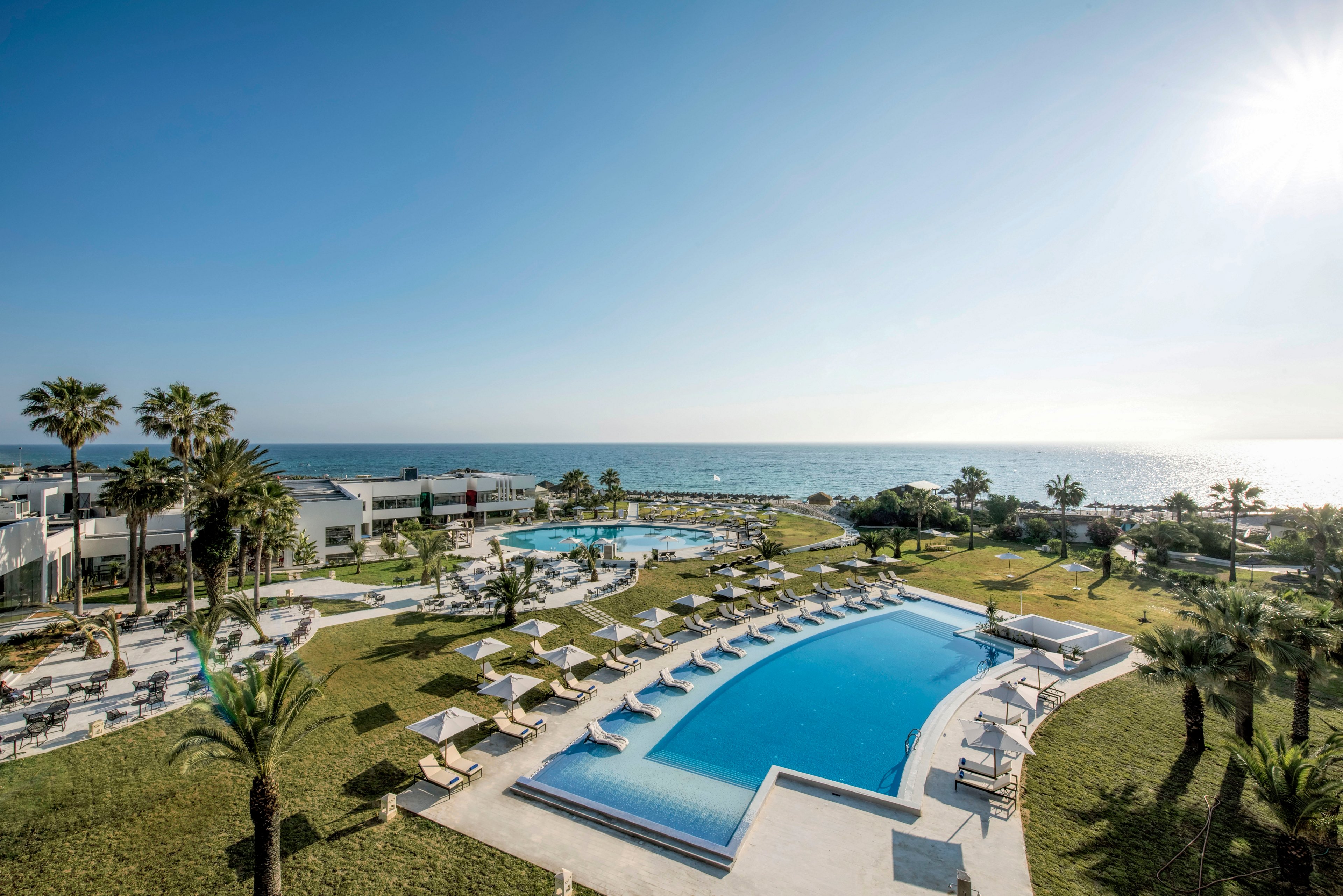 A large luxury resort features multiple outdoor swimming pools overlooking the sea. Sun loungers and umbrellas are neatly arranged around the main pool on manicured green lawns. Palm trees and landscaped gardens create a tropical atmosphere under a clear blue sky. Buildings and walkways frame the scene, emphasizing relaxation and vacation leisure by the waterfront.