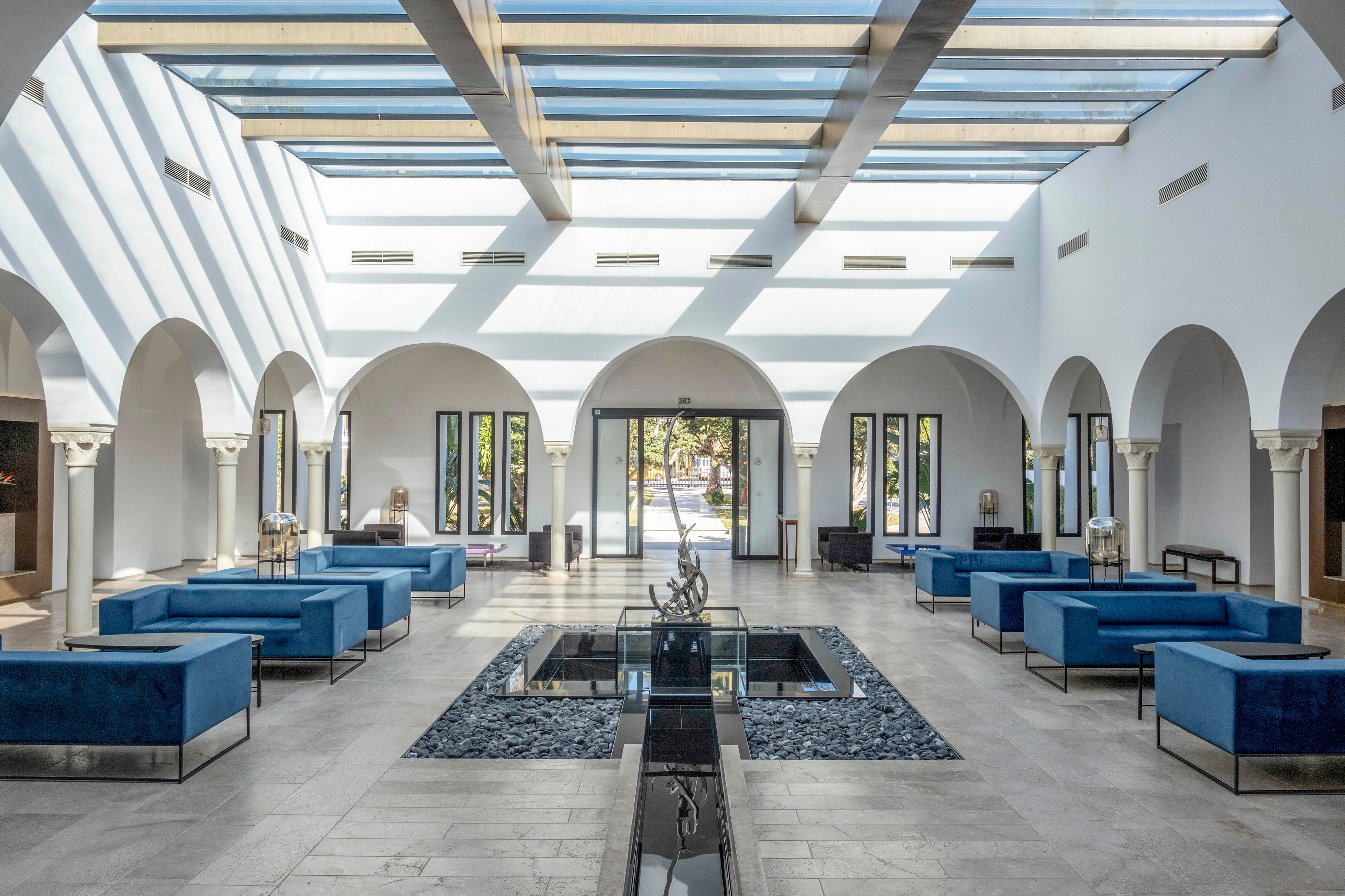 Spacious contemporary atrium lobby featuring multiple blue upholstered sofas arranged symmetrically. A central water feature with dark stone accents runs through the middle of the space. Tall white arches and columns frame the perimeter, leading to large glass doors that open to an outdoor area. A glass ceiling allows abundant natural light to cast shadows across the bright tiled floor, creating an airy and elegant atmosphere.