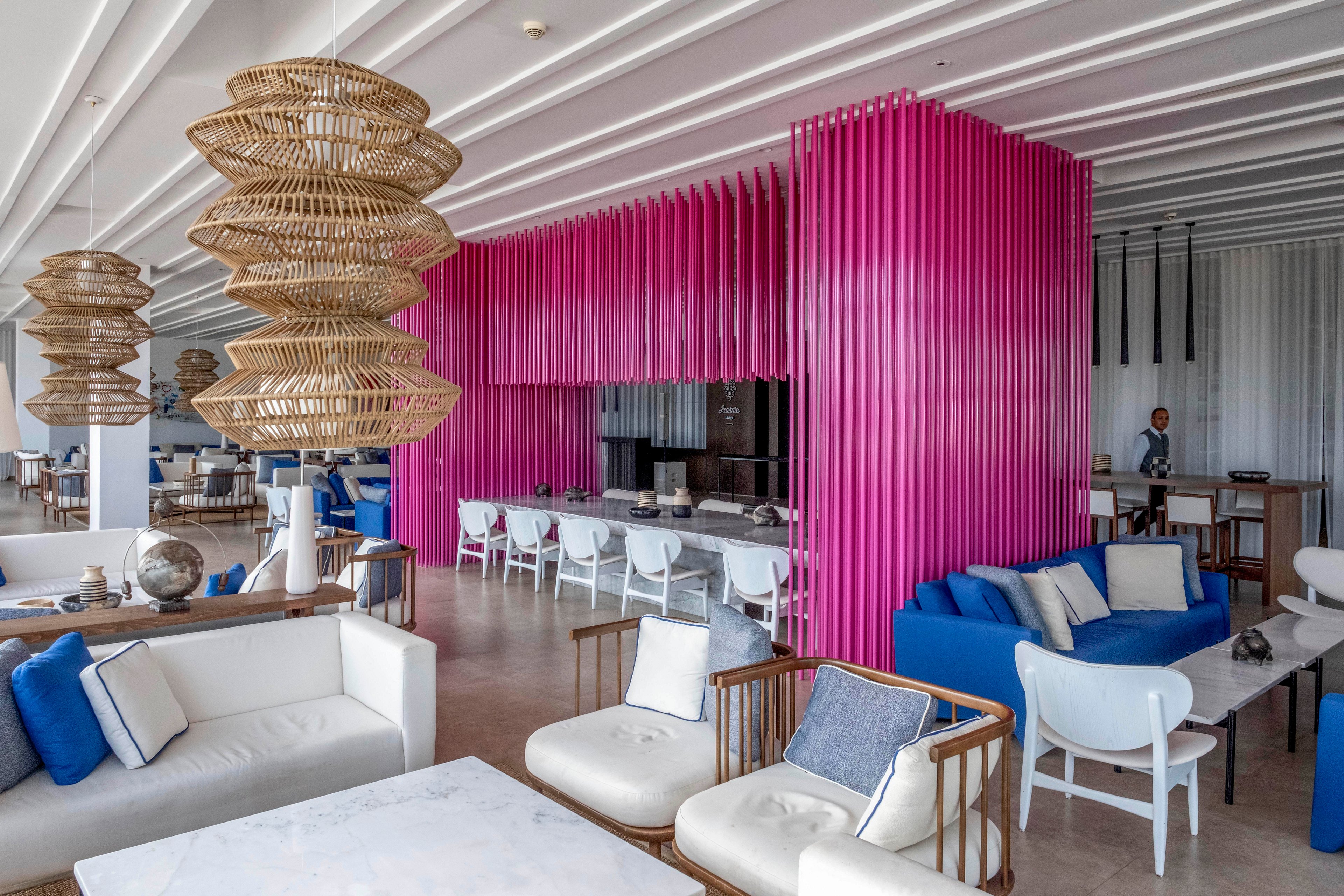 A contemporary resort lounge features white and blue seating arranged around low tables. A striking vertical pink partition structure defines the bar or dining area in the background. Woven rattan light fixtures hang from the ceiling, adding texture and warmth to the modern interior. A person stands near the back of the space, emphasizing the open, airy design.