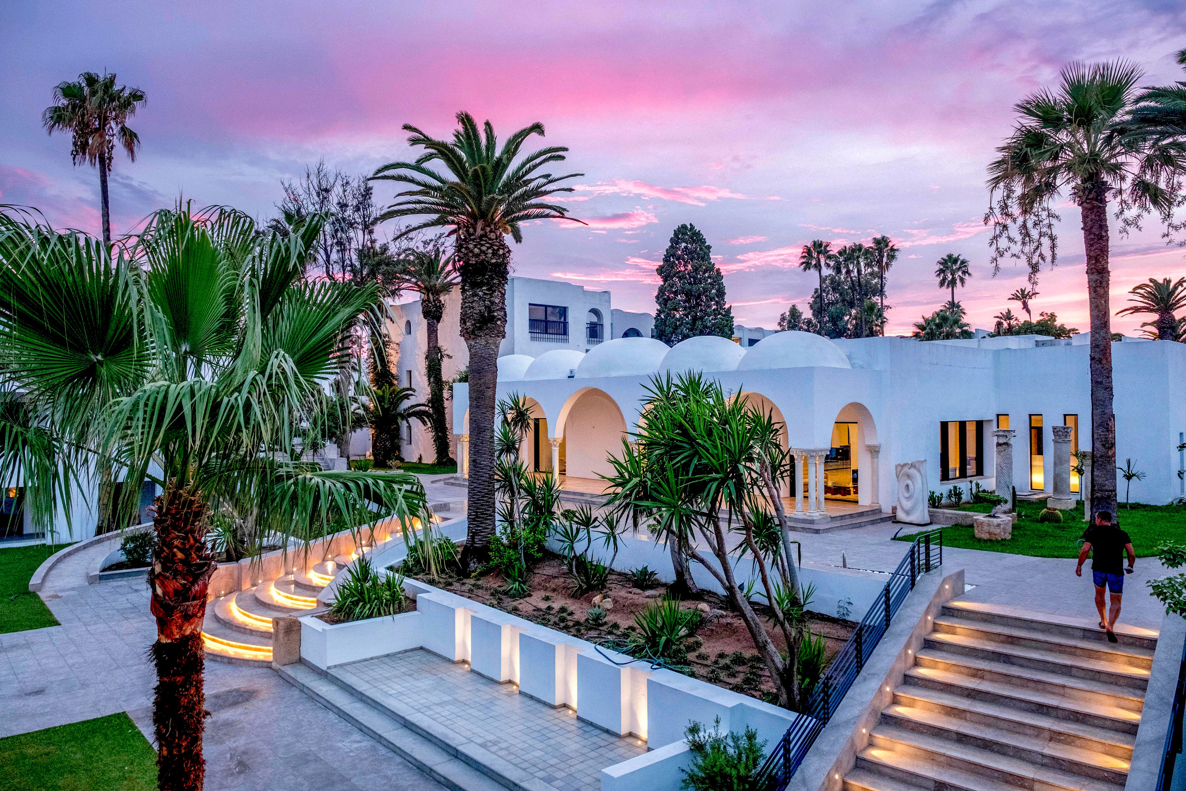 A modern white building with domed roofs is surrounded by lush palm trees and landscaped gardens. Wide staircases with illuminated steps lead up to the resort entrance, creating a welcoming pathway. The sky is filled with soft pink and purple hues, suggesting a tranquil sunset or sunrise. A single person walks along the steps, adding a sense of scale and relaxed vacation atmosphere.