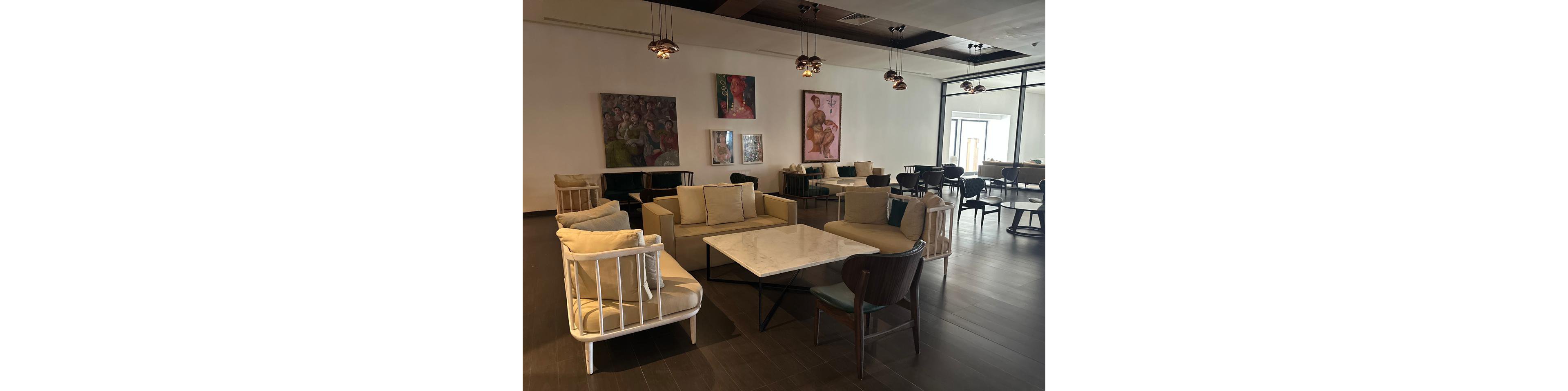 A modern lounge features beige sofas and armchairs arranged around a central marble coffee table. The space has dark wooden flooring and a high ceiling with pendant lights hanging above. Contemporary paintings decorate the back wall, adding color and artistic interest to the neutral interior. Large windows on the right allow natural light to brighten the seating area, creating a relaxed and inviting atmosphere.