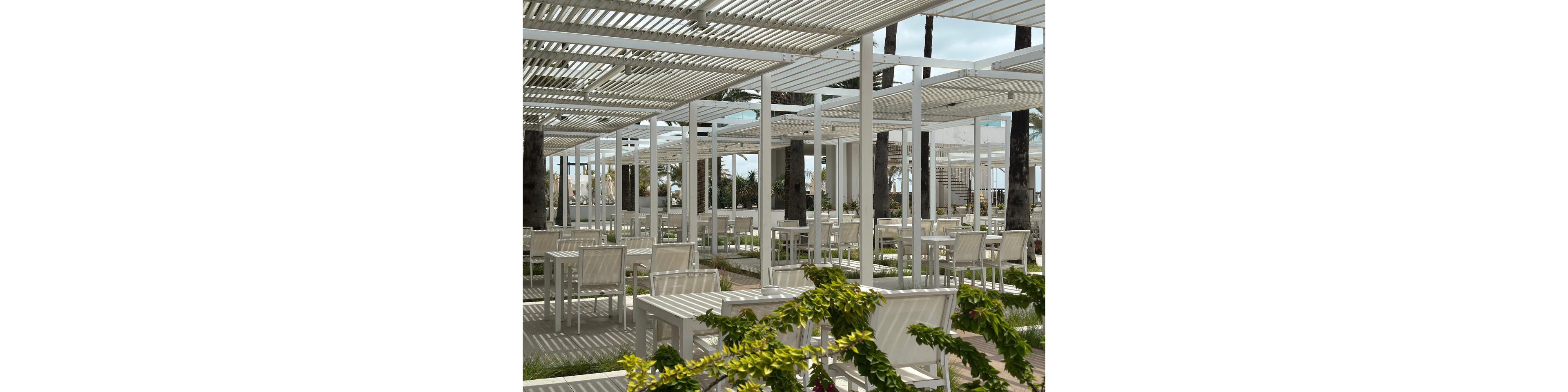 An empty outdoor dining terrace features rows of white tables and chairs beneath modern white pergolas. The space is open-air, with palm trees rising through the structure and glimpses of the sea and distant buildings in the background. Sunlight filters through the slatted roof, creating geometric shadows on the ground. Green plants and flowering vines add a touch of color to the otherwise neutral setting.