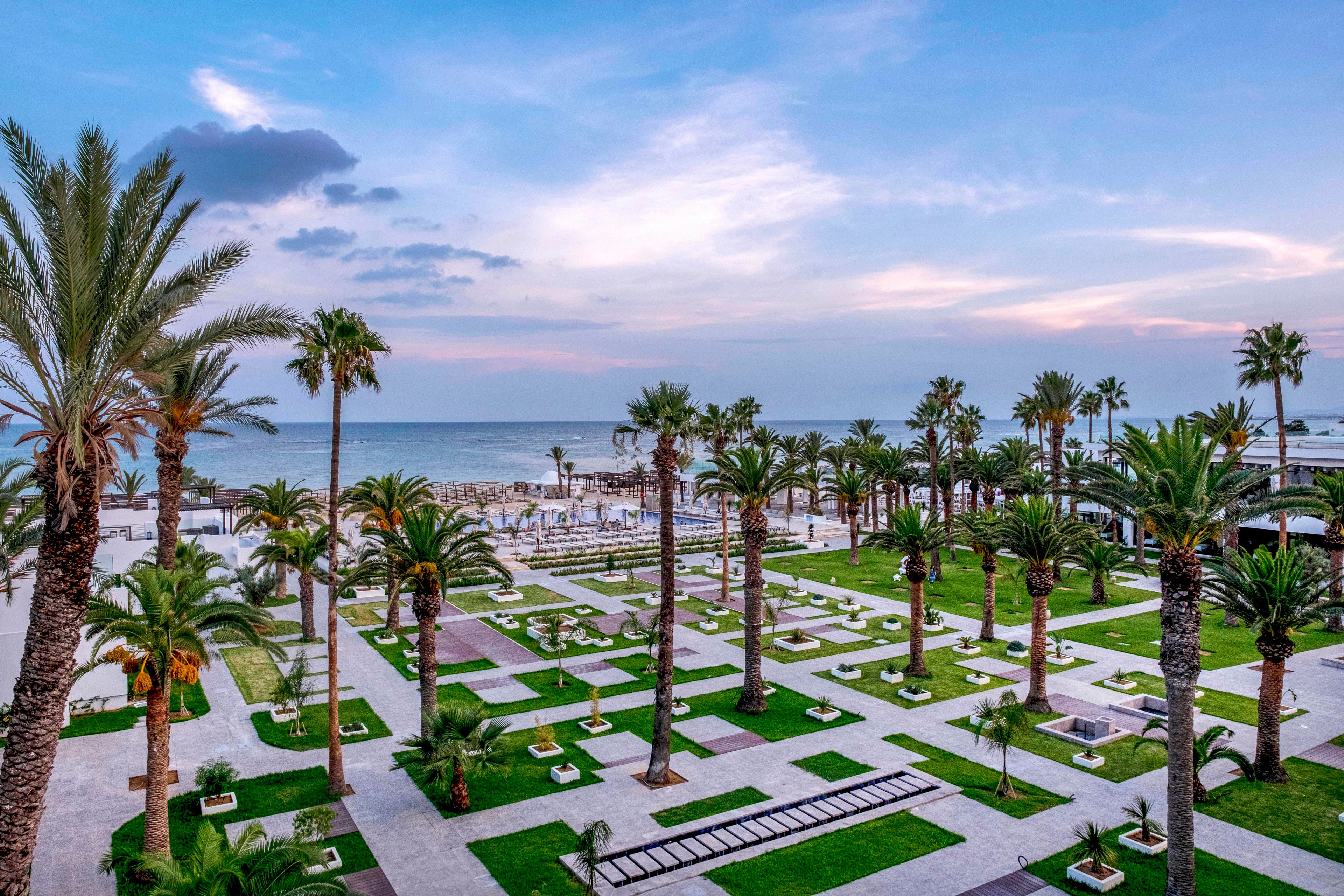A landscaped seaside resort garden stretches toward a calm ocean under a pastel evening sky. Tall palm trees line geometric lawns and paved walkways, creating a structured yet tropical atmosphere. Benches, low walls, and small planted sections add visual interest and places for relaxation. The soft sunset light and scattered clouds enhance the tranquil, vacation-like mood of the scene.