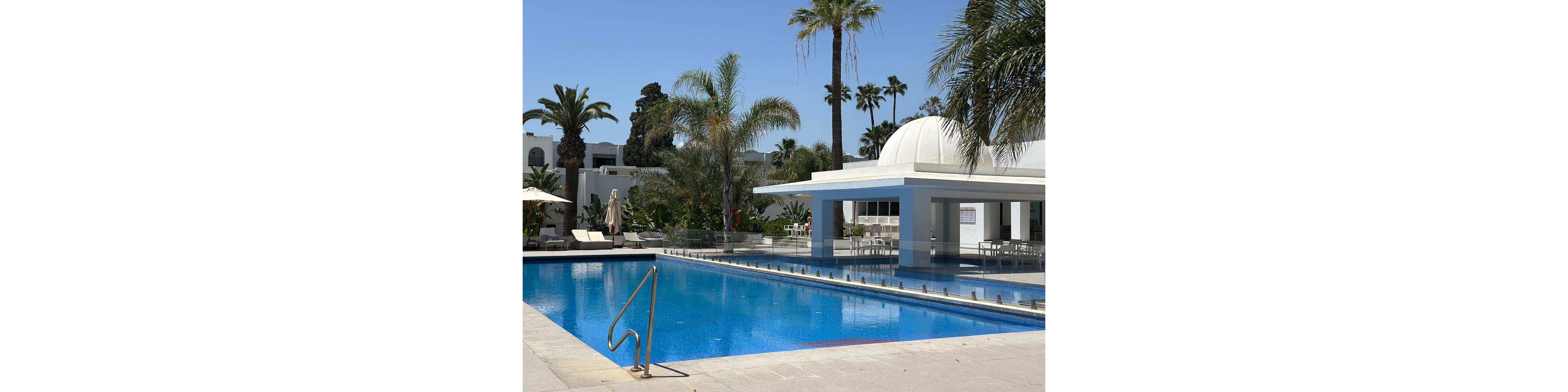 An outdoor swimming pool glistens under bright sunlight at a modern resort. White buildings with arches and a domed pool bar structure frame the water. Tall palm trees and lush greenery surround the poolside loungers and umbrellas. The clear blue sky and calm, empty seating area create a peaceful vacation atmosphere.