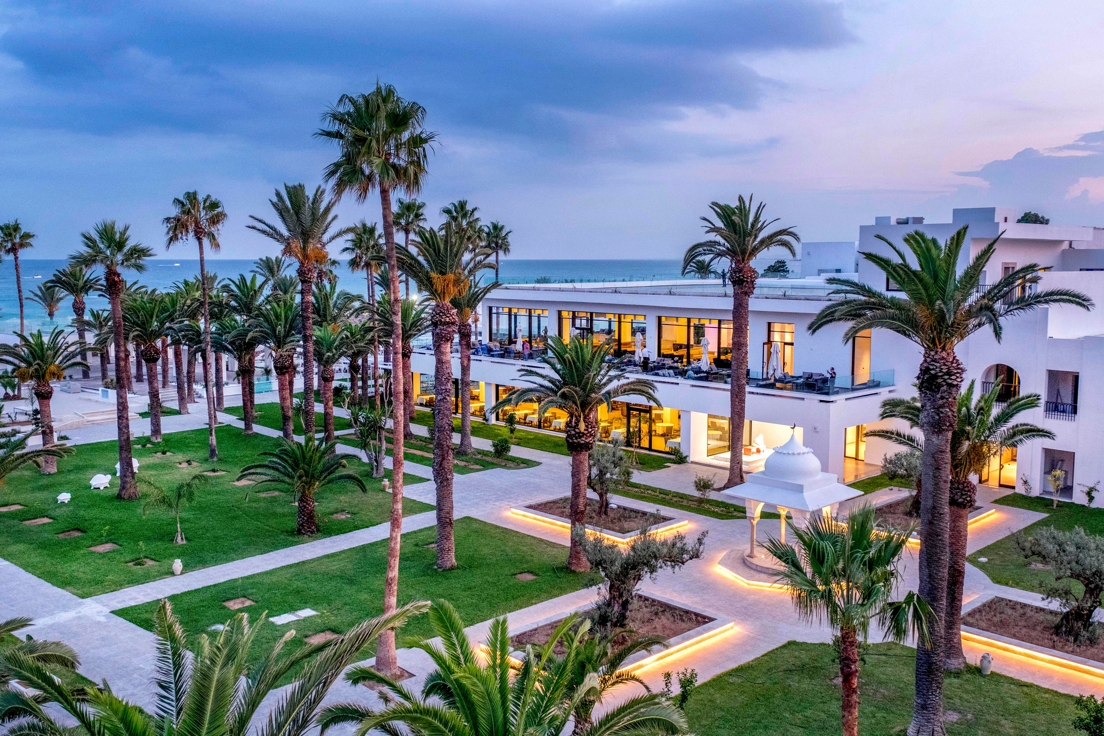 A contemporary white resort complex sits along a calm seaside shoreline at dusk. Tall palm trees and manicured green lawns surround the buildings, creating a tropical atmosphere. Warm interior lights glow through large windows, contrasting with the cool blue evening sky. Pathways and geometric landscaping elements lead toward the ocean, emphasizing the tranquil vacation setting.