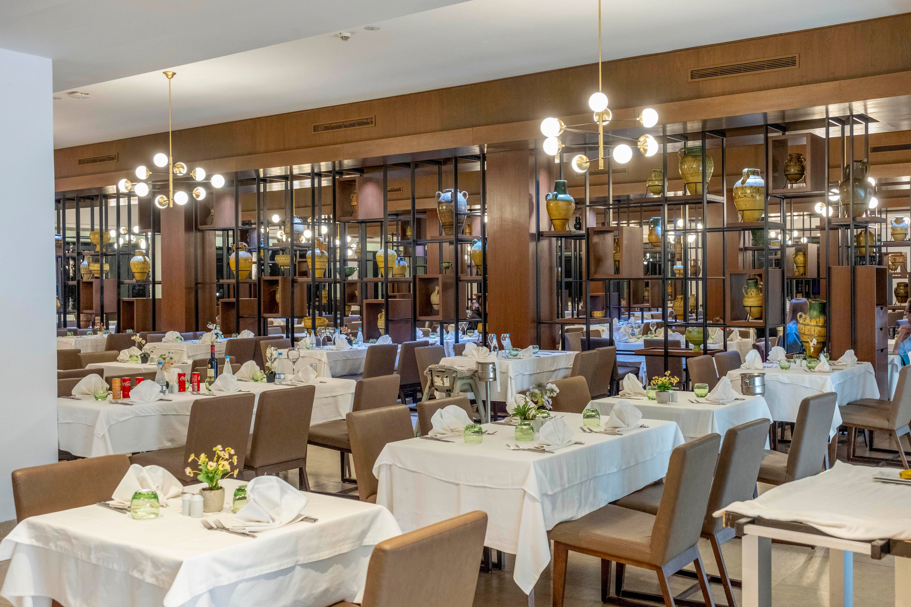 A spacious restaurant dining room is set with neatly arranged tables covered in white tablecloths. Brown upholstered chairs surround each table, which are decorated with folded napkins, glassware, and small floral centerpieces. Warm pendant lights hang from the ceiling, illuminating the space with a soft glow. A decorative wall of shelves with vases and objects forms the backdrop, creating a refined and contemporary atmosphere.