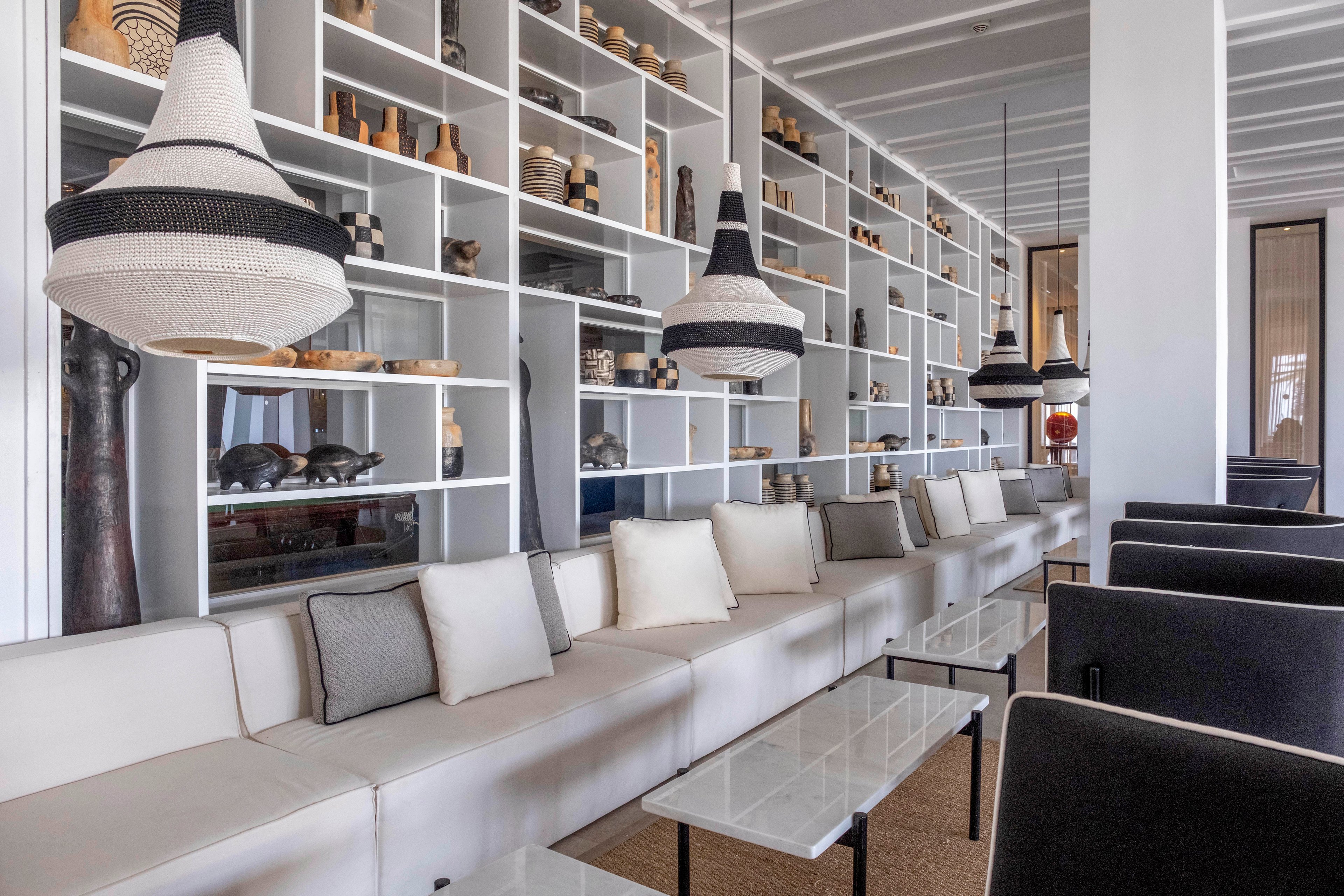 A contemporary lounge features long white sofas lined with neutral cushions facing sleek glass-topped tables. Behind the seating, a full-height wall of open shelving displays assorted vases, bowls, and sculptural objects. Large woven pendant lights hang from the ceiling, adding texture and visual interest to the minimalist space. The overall palette is soft and neutral, emphasizing clean lines and modern design.