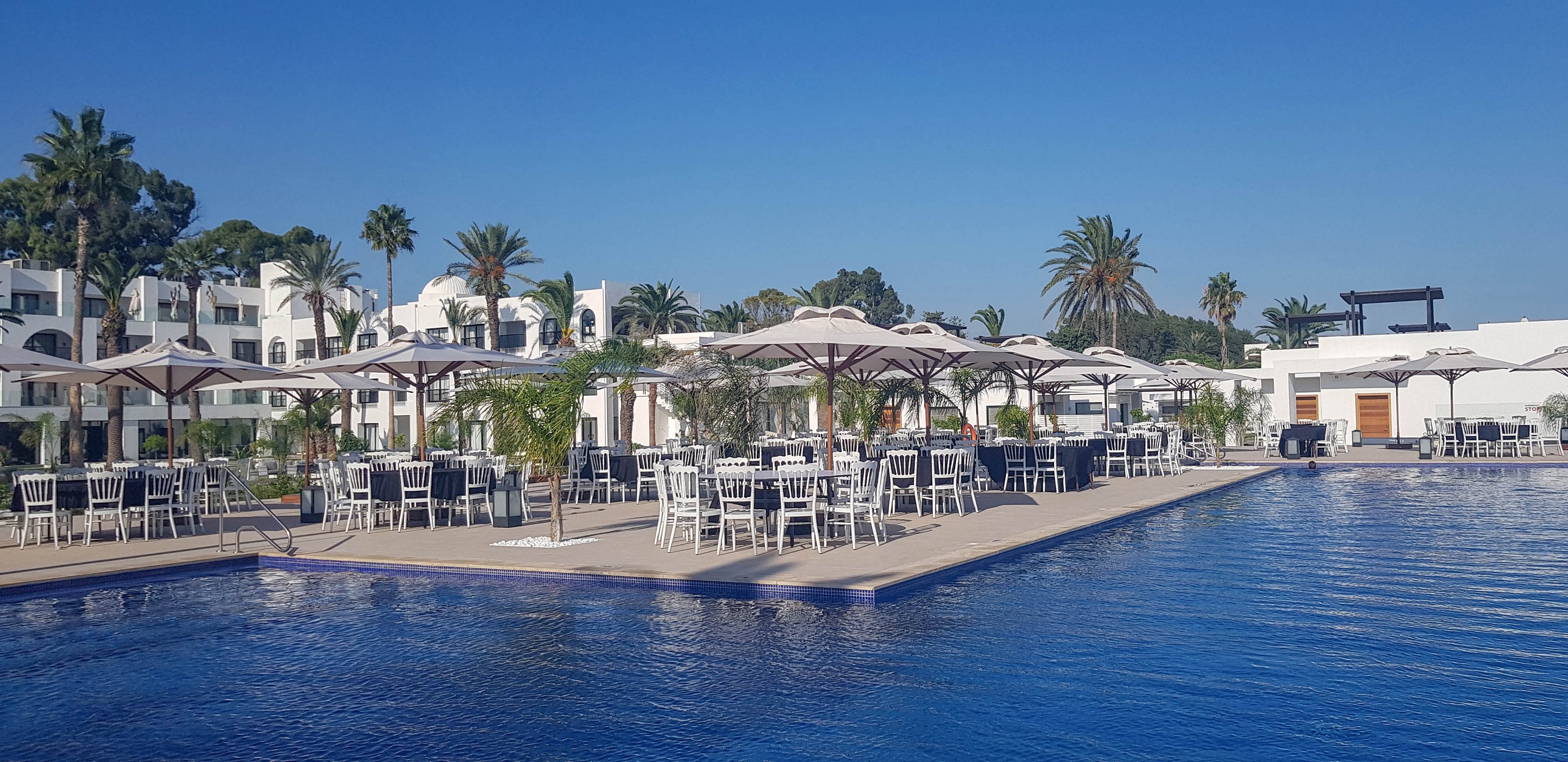 An expansive outdoor dining area is set up beside a large blue swimming pool at a resort. White tables and chairs are arranged neatly under beige umbrellas, suggesting preparation for an event or meal service. White low-rise buildings with balconies and arched windows frame the background, surrounded by tall palm trees and lush greenery. The clear blue sky and calm water create a relaxed, vacation-like atmosphere.
