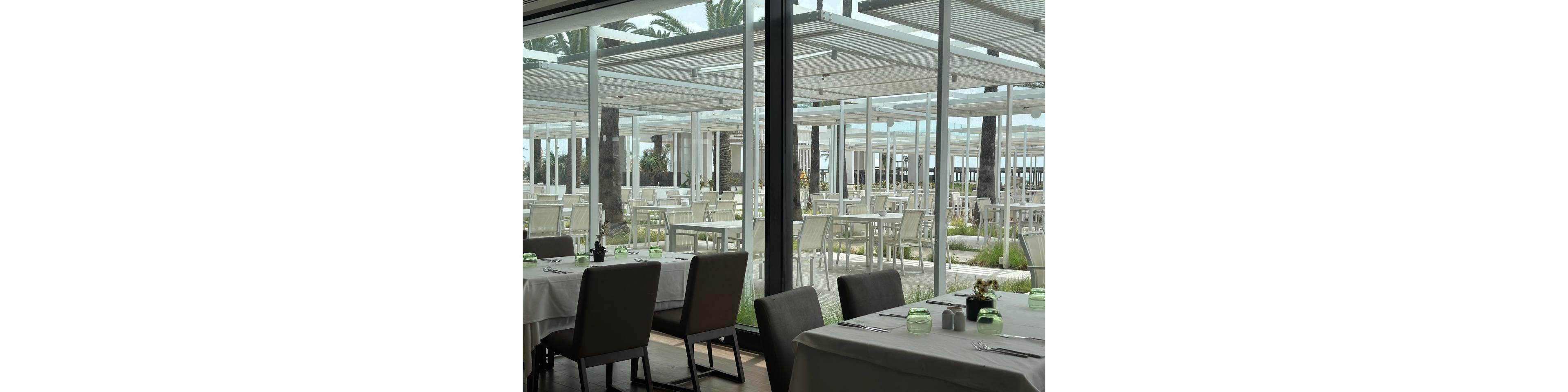 A contemporary restaurant dining area overlooks an expansive outdoor terrace filled with empty white tables and chairs. The scene is framed by large glass windows and sleek metal pergolas that provide shade. Soft neutral tones dominate the interior, accented by green glass candle holders on the tables. Palm trees and glimpses of the sea in the background suggest a coastal resort setting.