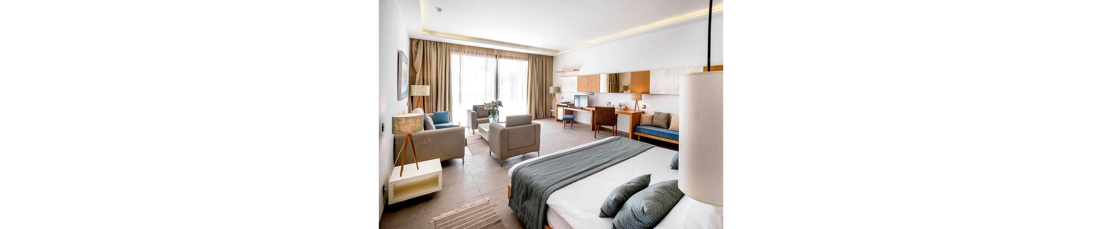 Spacious contemporary hotel room featuring a large bed with blue-gray bedding in the foreground. A cozy seating area with armchairs and a sofa is arranged near wide floor-to-ceiling windows covered by sheer curtains. A long wooden desk with chairs, lamps, and decorative wall panels lines the opposite wall. Neutral tones, natural light, and minimalist decor create a calm, inviting atmosphere suitable for relaxation or work.