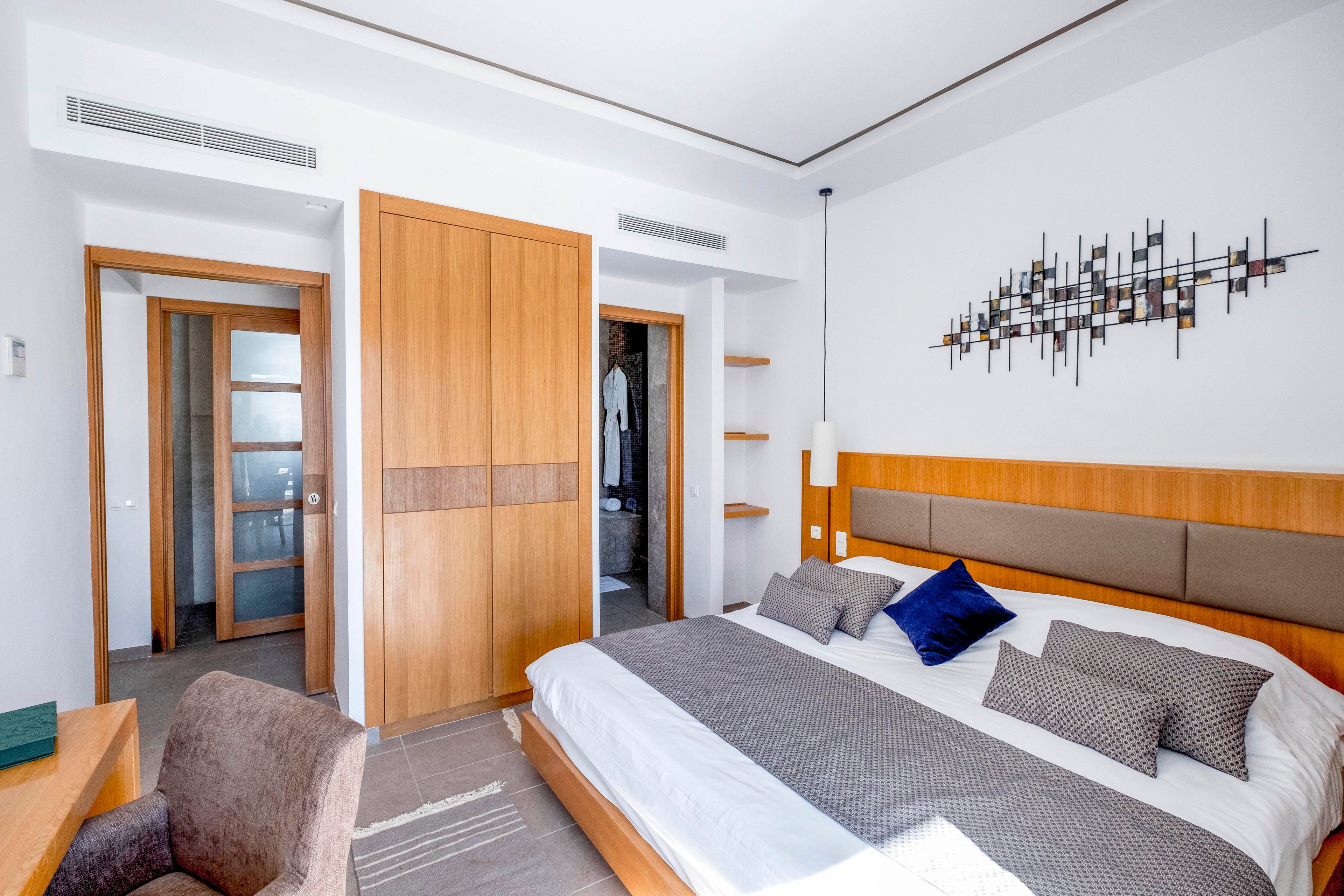 A contemporary hotel-style bedroom features a large bed with gray and white bedding and accent pillows. Warm wooden elements dominate the space, including built-in wardrobes, doors, and a headboard. A small desk and upholstered chair sit in the corner, while a geometric wall sculpture decorates the white wall above the bed. Soft natural light brightens the clean, minimalist interior.