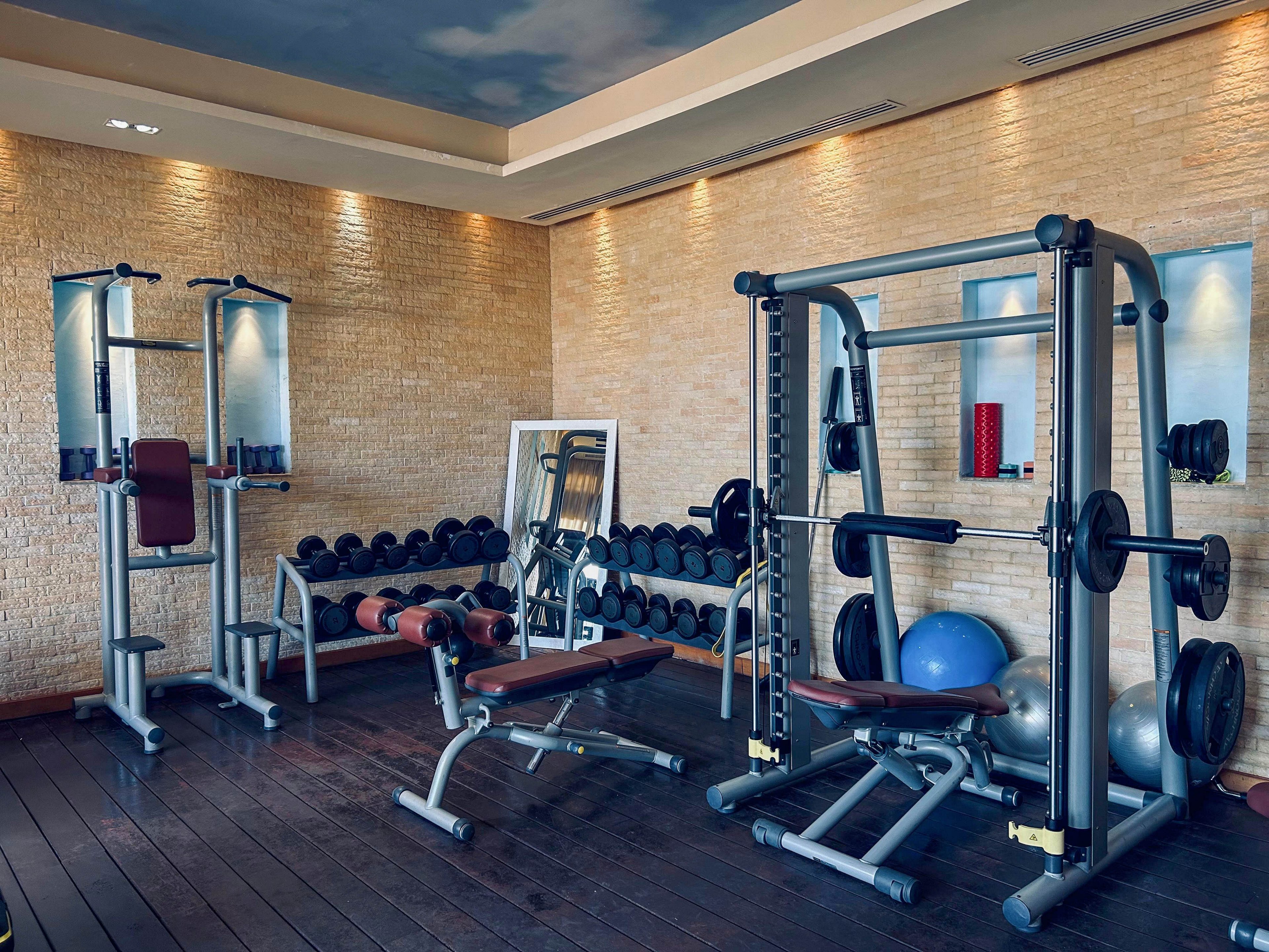 A compact fitness room features several strength training machines and free weights neatly arranged along a textured beige wall. Dumbbells are lined up on racks beside benches and a multi-station weight machine. Exercise balls and other accessories add variety to the workout options. Recessed ceiling lights and a painted sky ceiling create a bright, inviting atmosphere on the dark wooden floor.