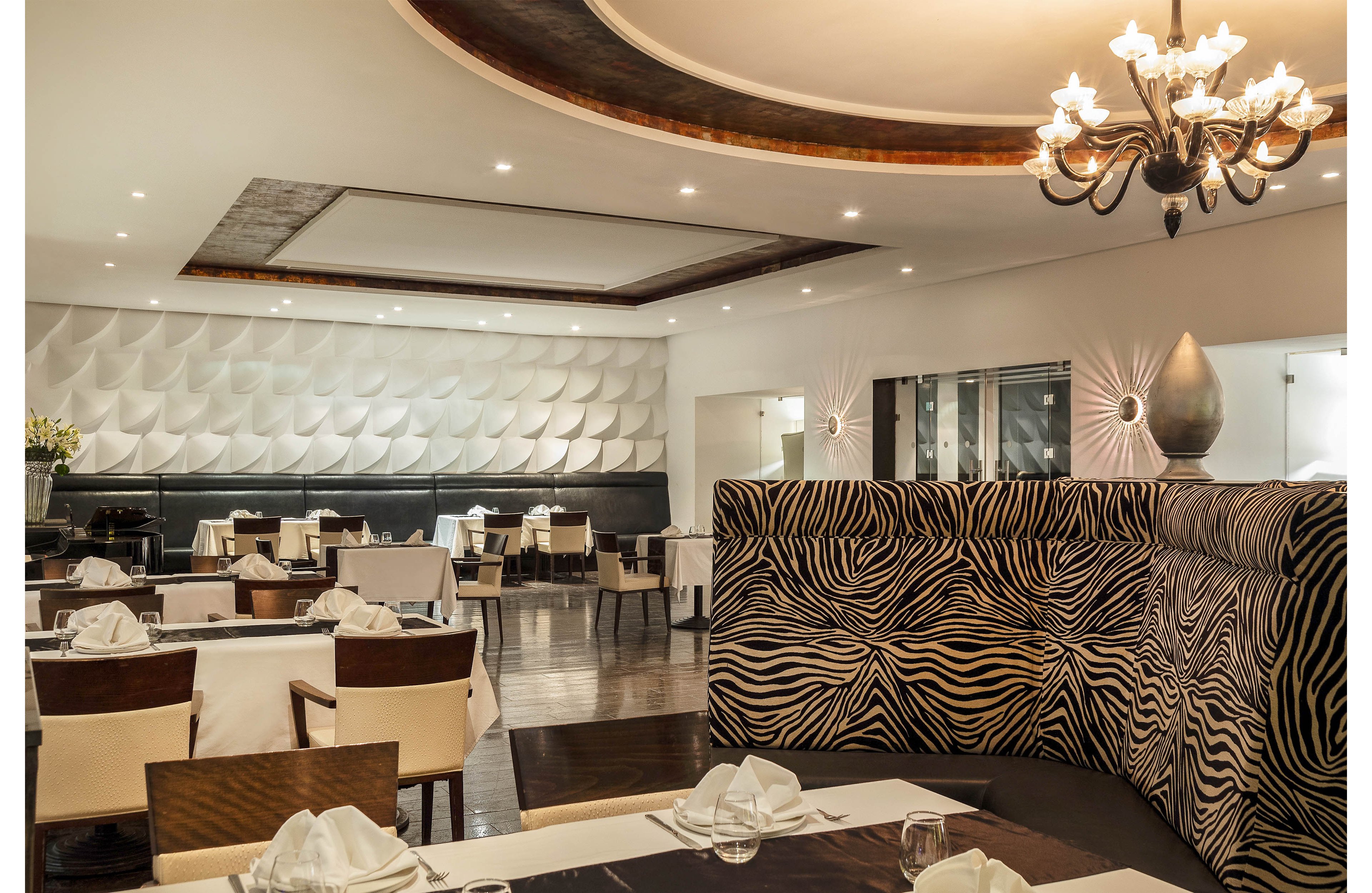 A contemporary restaurant dining room features neatly set tables with white tablecloths and folded napkins. A bold zebra-patterned booth divides the seating area, adding contrast to the neutral decor. Soft ceiling lighting and a decorative chandelier create a warm, inviting atmosphere. The space appears empty of people, emphasizing the clean, elegant design and stylish wall textures.