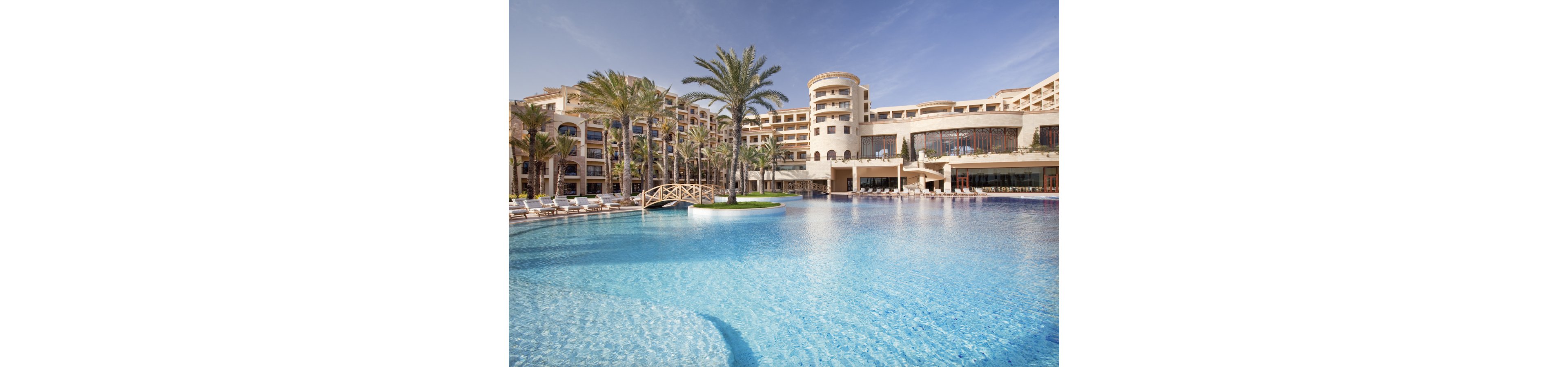 A large luxury resort hotel surrounds an expansive outdoor swimming pool under a clear blue sky. Palm trees and sun loungers line the poolside, creating a relaxing vacation atmosphere. A small decorative bridge crosses part of the pool, adding architectural interest to the scene. The bright sunlight and reflections in the clear water emphasize a warm, tranquil holiday setting.