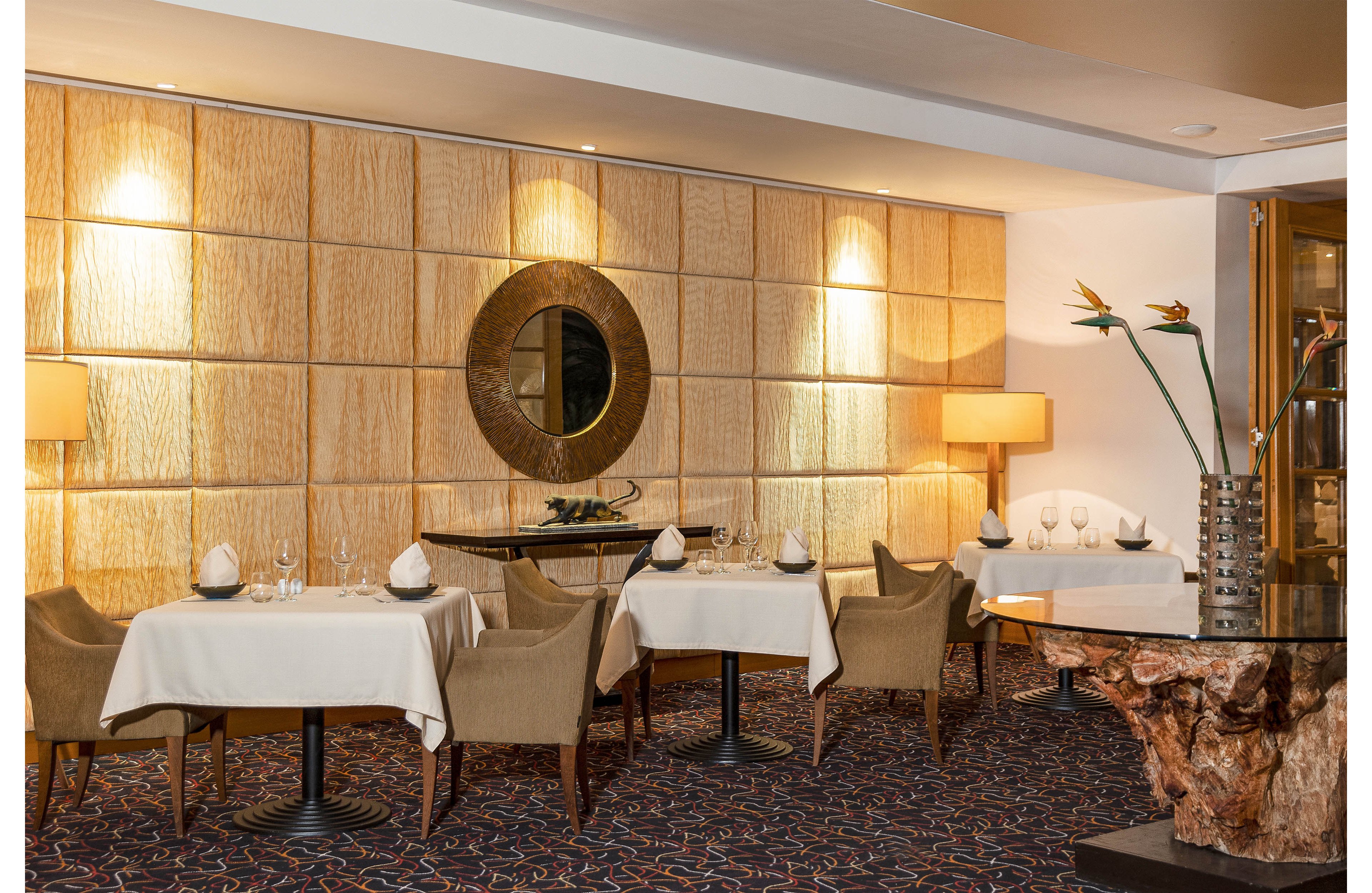 Upscale restaurant dining area with neatly set tables and upholstered chairs is shown. White tablecloths, folded napkins, and arranged glassware create a refined fine dining atmosphere. Warm wall sconces illuminate a textured wall featuring a large circular mirror as a focal point. A sculptural wooden console with tall vases and flowers adds a touch of natural decor to the sophisticated interior.