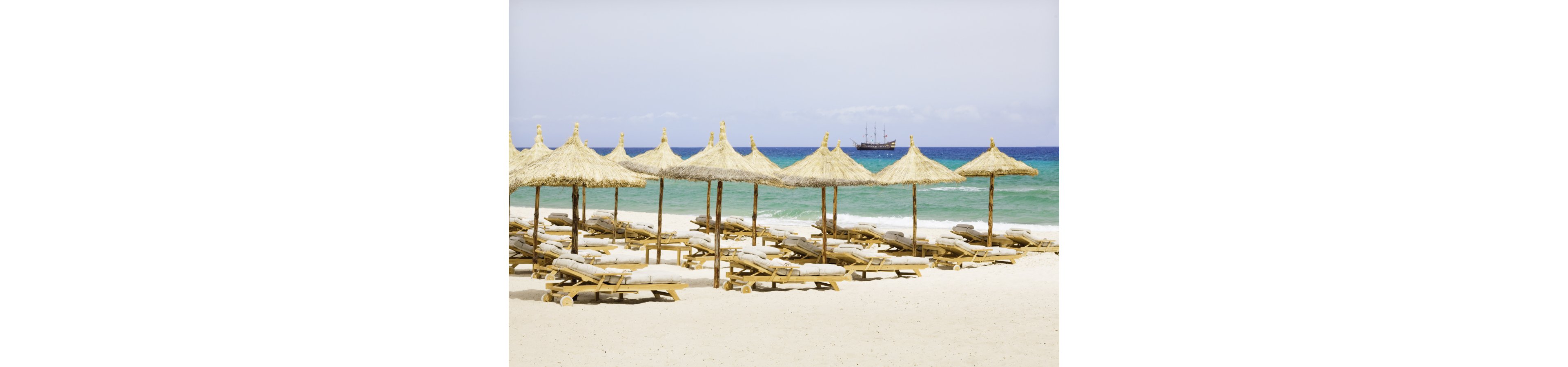 A row of wooden sun loungers with cushions sits beneath straw umbrellas on a bright sandy beach. The turquoise sea stretches out to the horizon under a pale blue sky. In the distance, a ship sails across the calm water. The scene feels peaceful and inviting, suggesting a quiet seaside resort or vacation destination.
