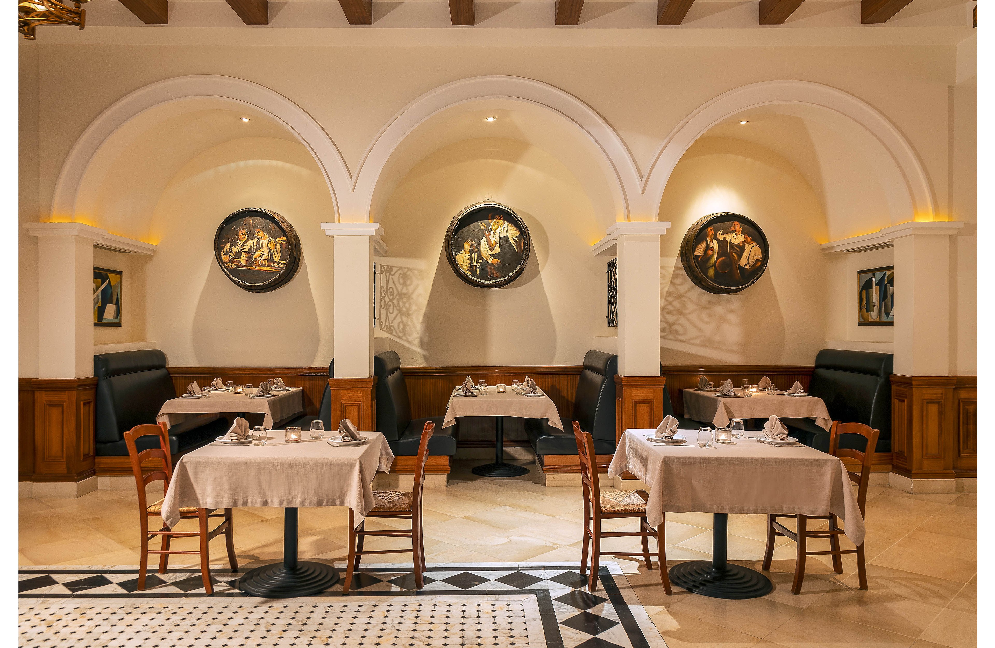 An upscale restaurant interior features neatly set tables arranged before three arched alcoves. Each alcove contains a black leather booth and framed artwork illuminated by warm lighting. White tablecloths, folded napkins, and glassware create a refined dining atmosphere. The tiled floor with a black and white geometric border adds contrast to the cream-colored walls and ceiling beams.