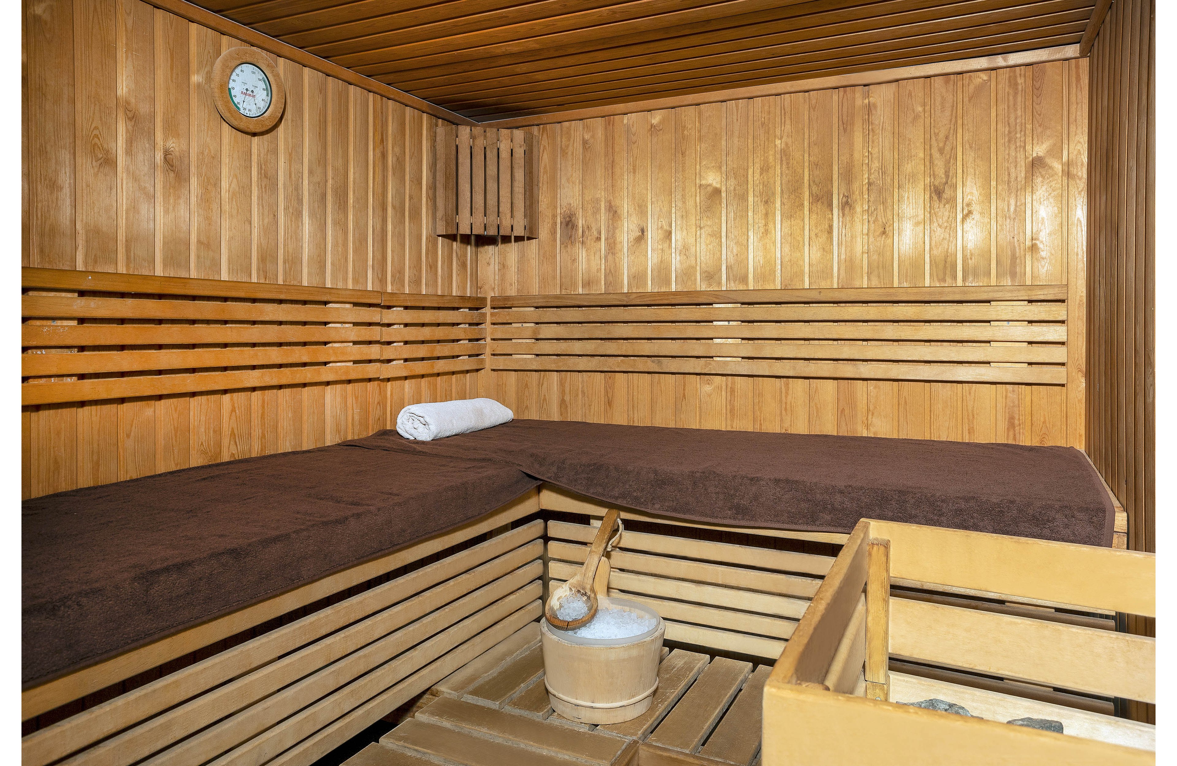 A clean wooden sauna interior features wide tiered benches covered with dark brown mats. A rolled white towel rests neatly on the upper bench, suggesting readiness for relaxation. In the foreground, a wooden bucket with a ladle sits beside a container of sauna stones. The walls and ceiling are lined with warm-toned wood panels, and a small round wall clock is visible, emphasizing a calm, orderly atmosphere.
