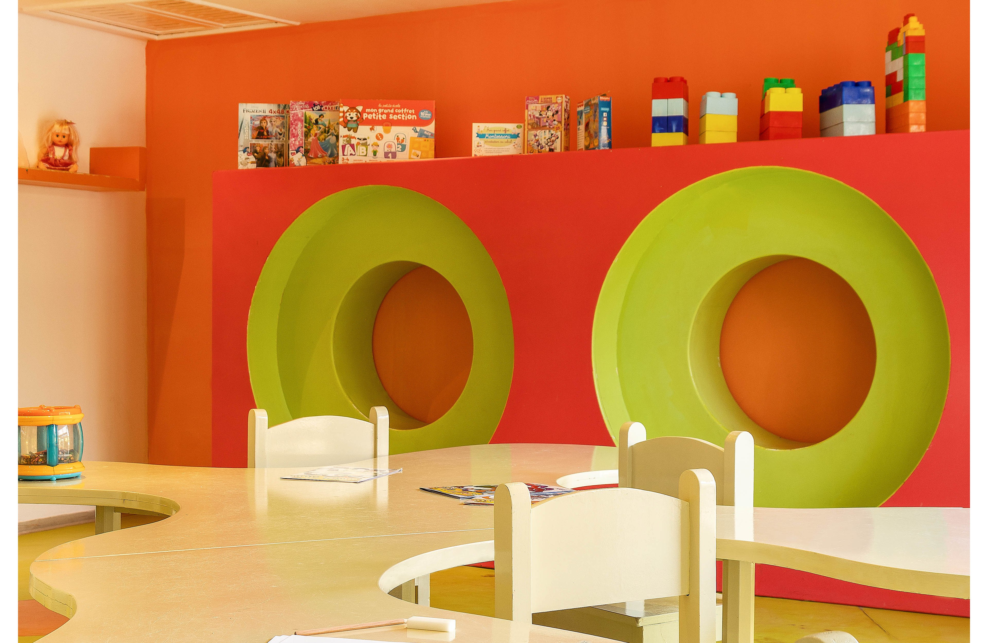 A bright children's playroom features curved tables and small white chairs arranged in the foreground. The back wall is painted orange with large green circular cutouts that create playful shelf niches. Toys, books, and large building blocks are neatly displayed along the top of the wall. The overall atmosphere is cheerful and creative, designed to engage young children in play and learning.