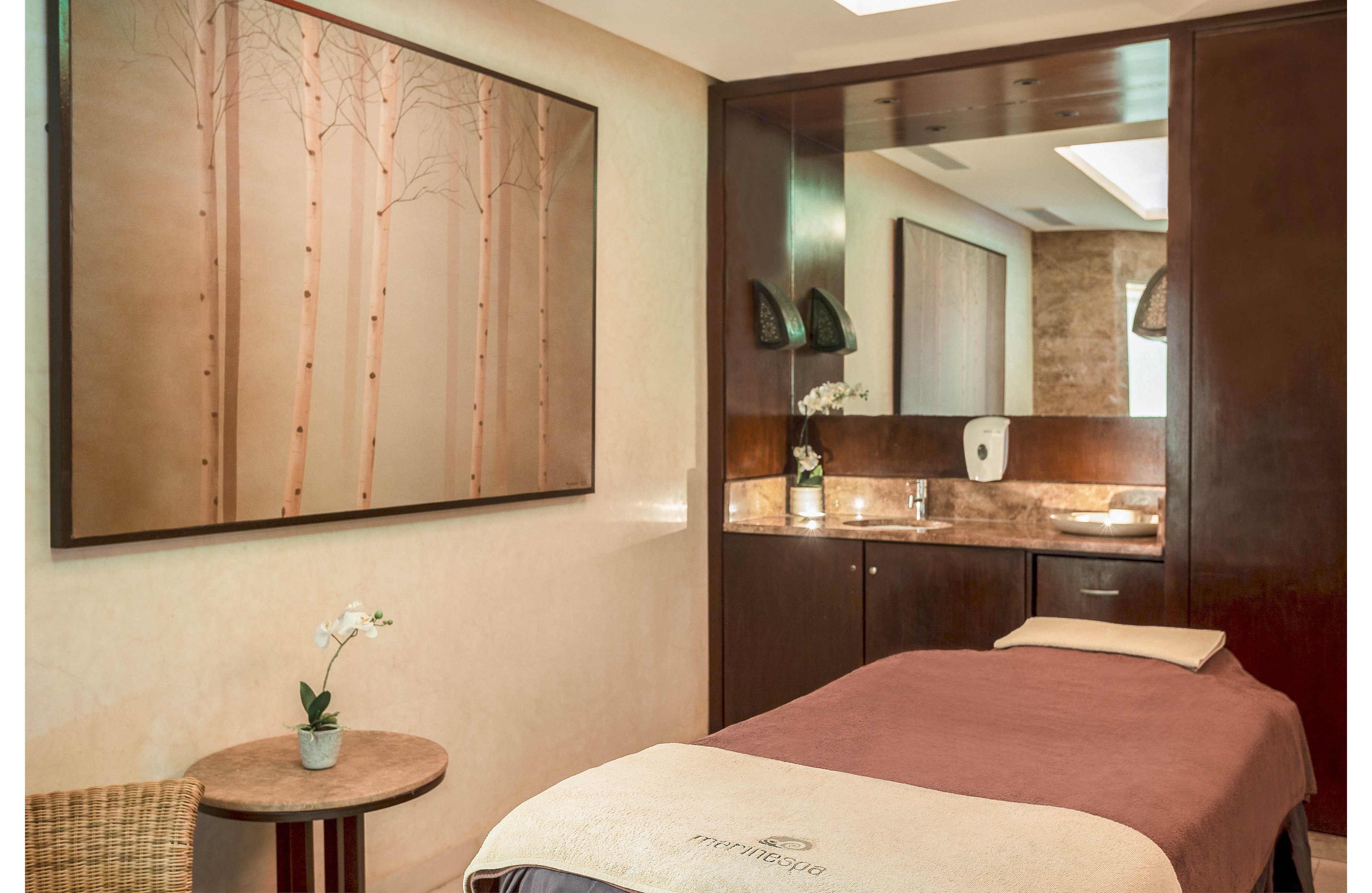 A tranquil spa treatment room features a neatly made massage table with a soft brown blanket. The space includes a wooden side table holding a small potted orchid, adding a touch of nature. Warm lighting reflects off a countertop with a sink and mirror, creating a cozy ambiance. A large wall art piece with tree motifs enhances the calming, minimalist decor.