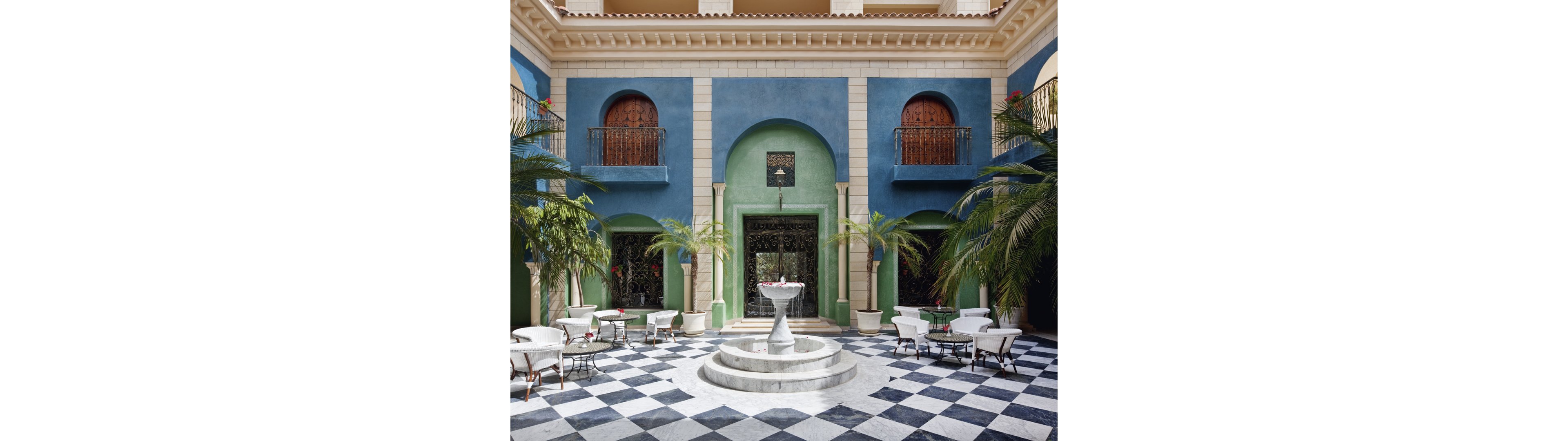 An elegant courtyard features a white marble fountain at its center, surrounded by checkerboard black and white floor tiles. Blue and green walls with arched doorways and wrought iron details create a Mediterranean or Moorish-inspired setting. Potted palm trees and greenery soften the architecture and frame the seating areas. White chairs and small round tables suggest an outdoor café or hotel patio, evoking a relaxed and refined atmosphere.