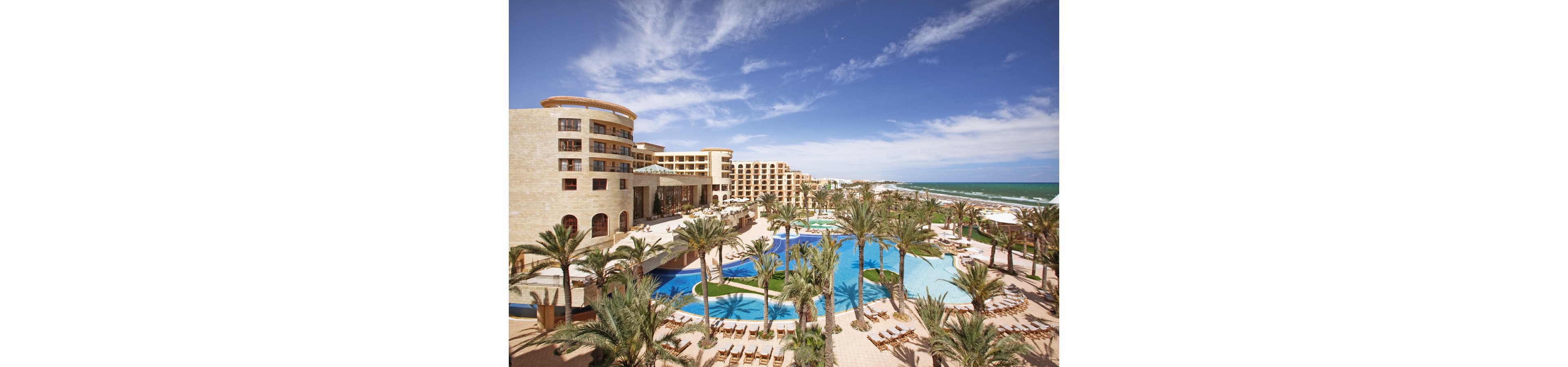 A large luxury resort complex overlooks a sandy beachfront and turquoise sea. Multiple curving swimming pools are surrounded by tall palm trees and rows of sun loungers. The hotel buildings feature warm-toned stone facades and balconies facing the ocean. Clear blue skies and bright sunlight create a relaxing vacation atmosphere.