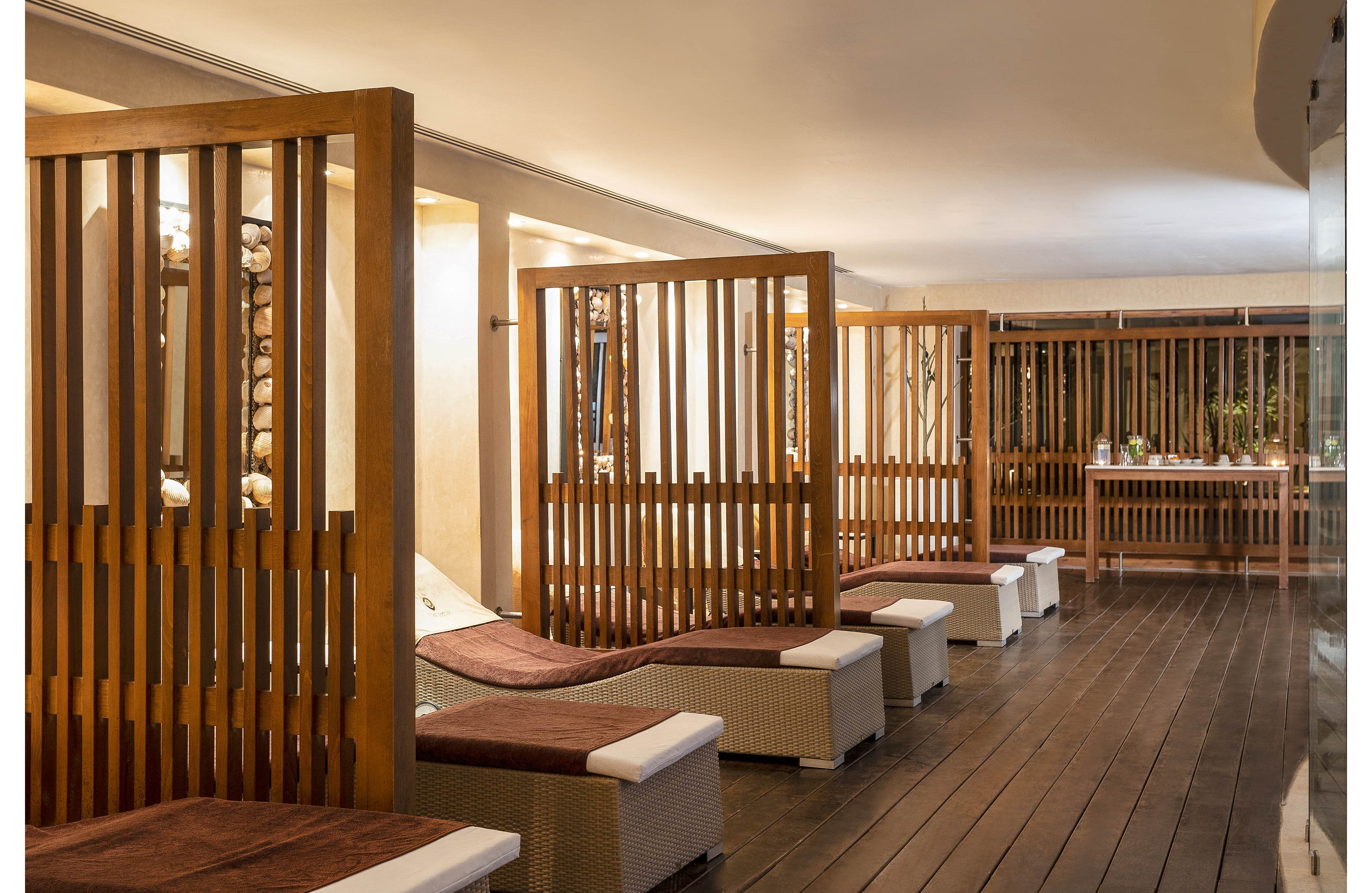 A contemporary spa relaxation area features a row of cushioned lounge chairs separated by tall wooden slatted partitions. The space has warm ambient lighting that highlights the natural wood tones and dark plank flooring. Soft brown and white upholstery adds comfort and contrast to the minimalist design. In the background, a counter with decor elements enhances the tranquil, resort-like atmosphere.
