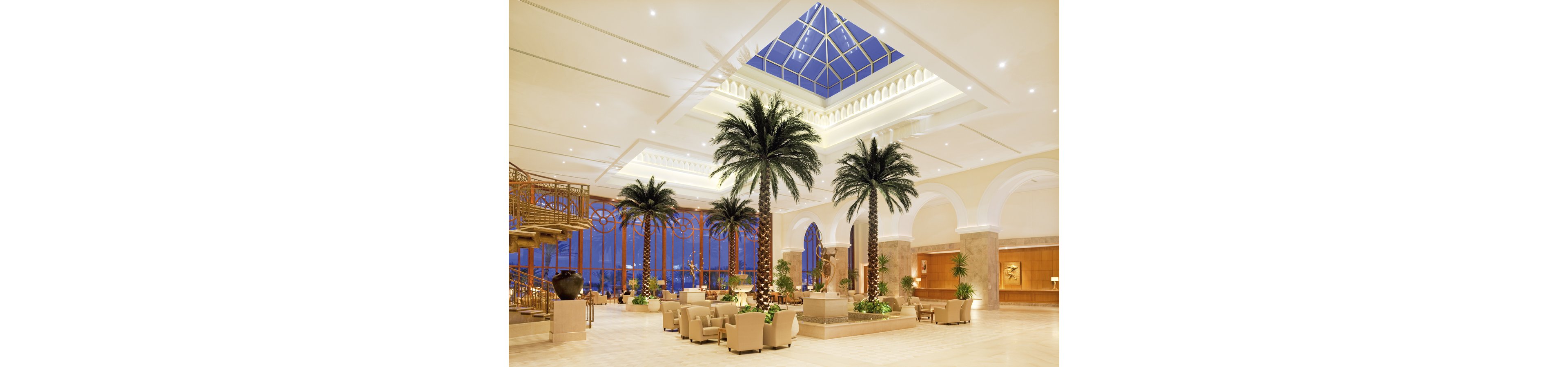 Spacious hotel lobby featuring tall indoor palm trees and elegant seating areas. Large cream-colored armchairs and small tables are arranged for guests to relax and socialize. A dramatic glass skylight and expansive windows reveal the evening sky outside. Warm lighting, marble floors, and decorative staircases create a sophisticated, resort-like atmosphere.