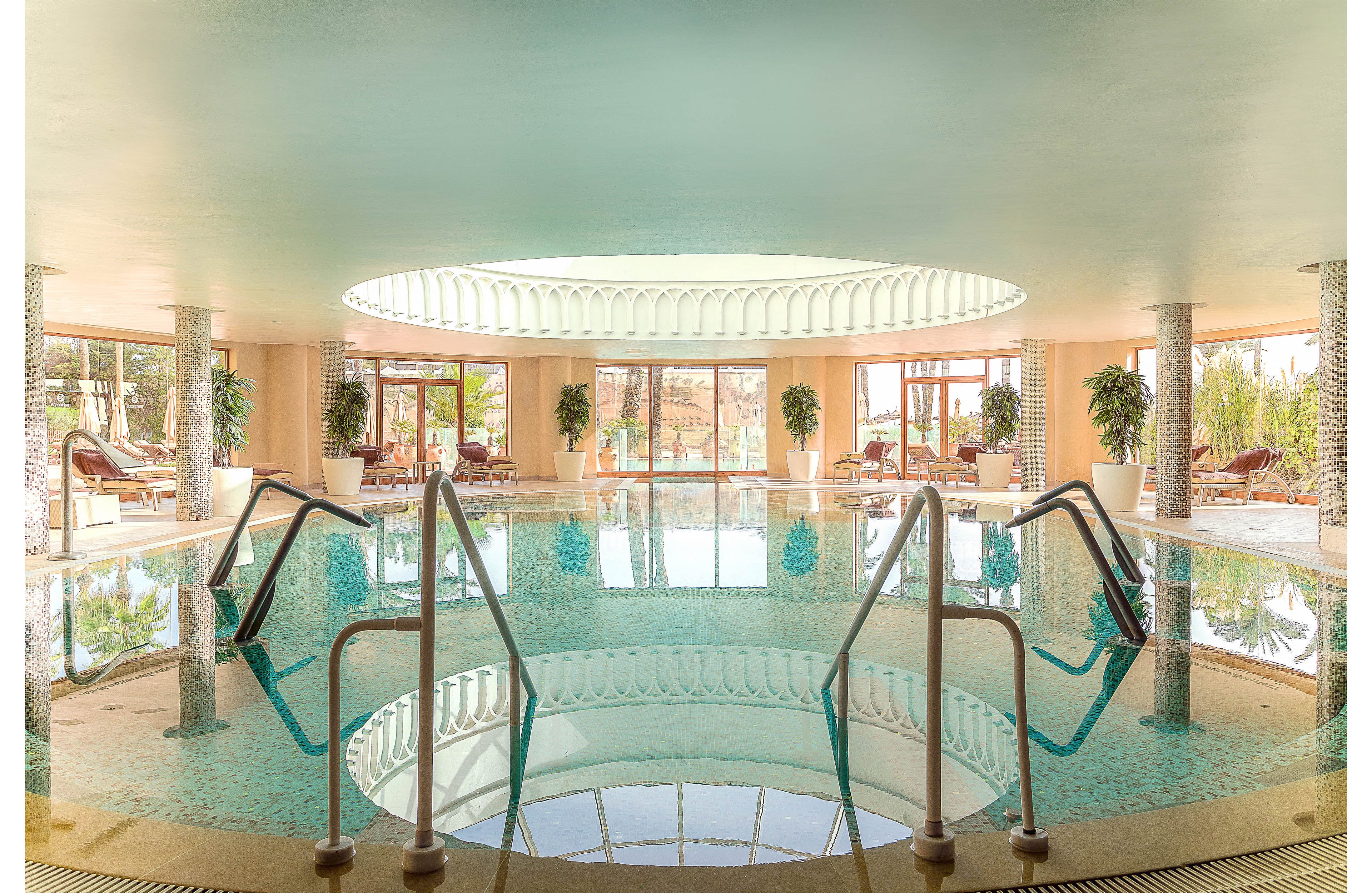 A circular indoor swimming pool sits beneath a large skylight, creating a bright and tranquil spa environment. Stainless steel handrails lead into the clear water, which reflects the ceiling and surrounding architecture. Sun loungers and potted plants line the perimeter, suggesting a relaxing wellness or resort setting. Large windows open to an outdoor view, enhancing the airy and serene atmosphere.