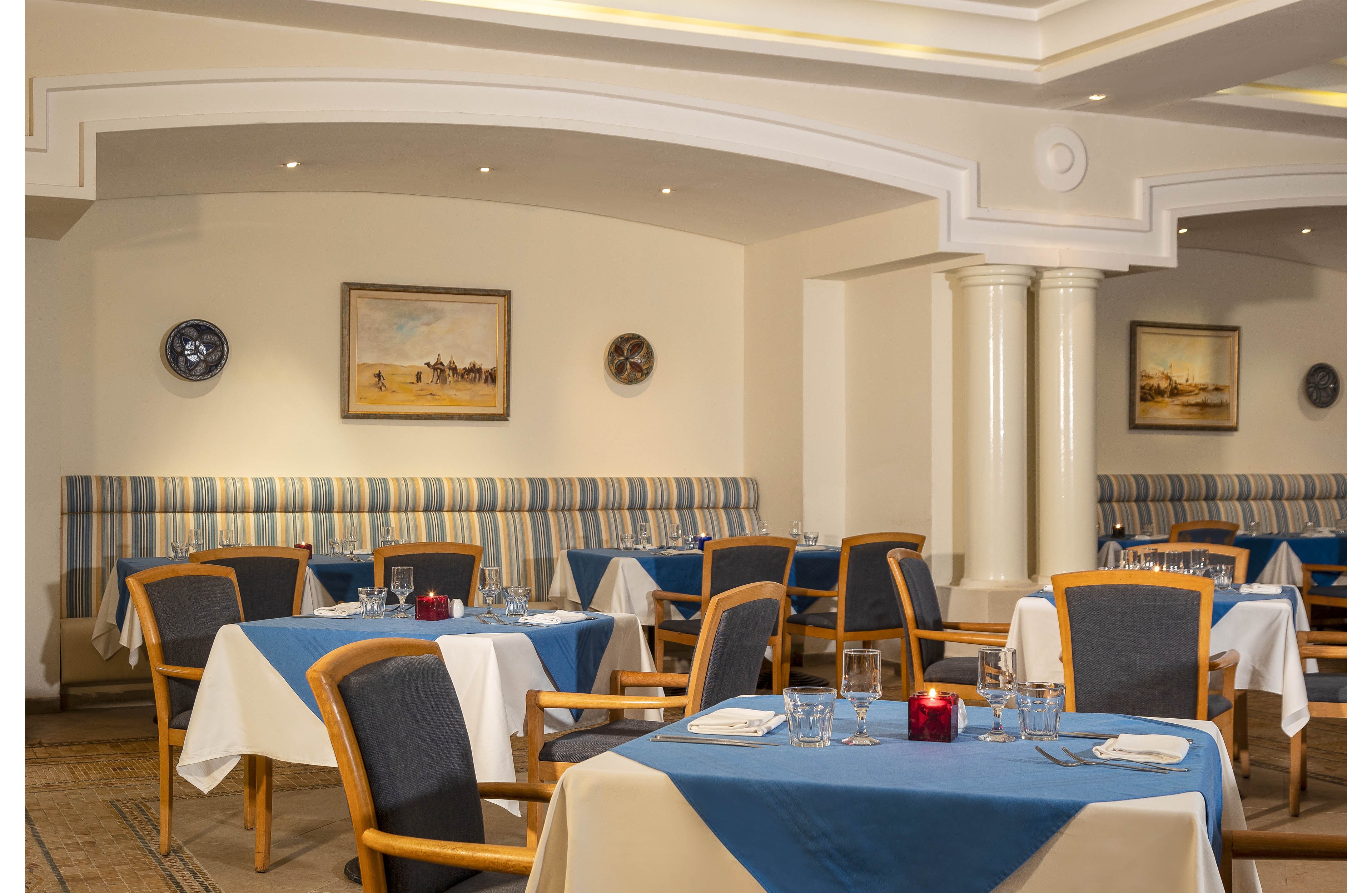 A neatly arranged restaurant dining room features several tables covered with white cloths and blue table runners. Wooden chairs with cushioned backs surround the tables, which are set with glassware, cutlery, and folded napkins. The space has a warm, neutral color palette with striped banquette seating along the wall and framed artwork as decoration. Soft ceiling lighting and white columns contribute to the refined, inviting atmosphere of the empty dining area.