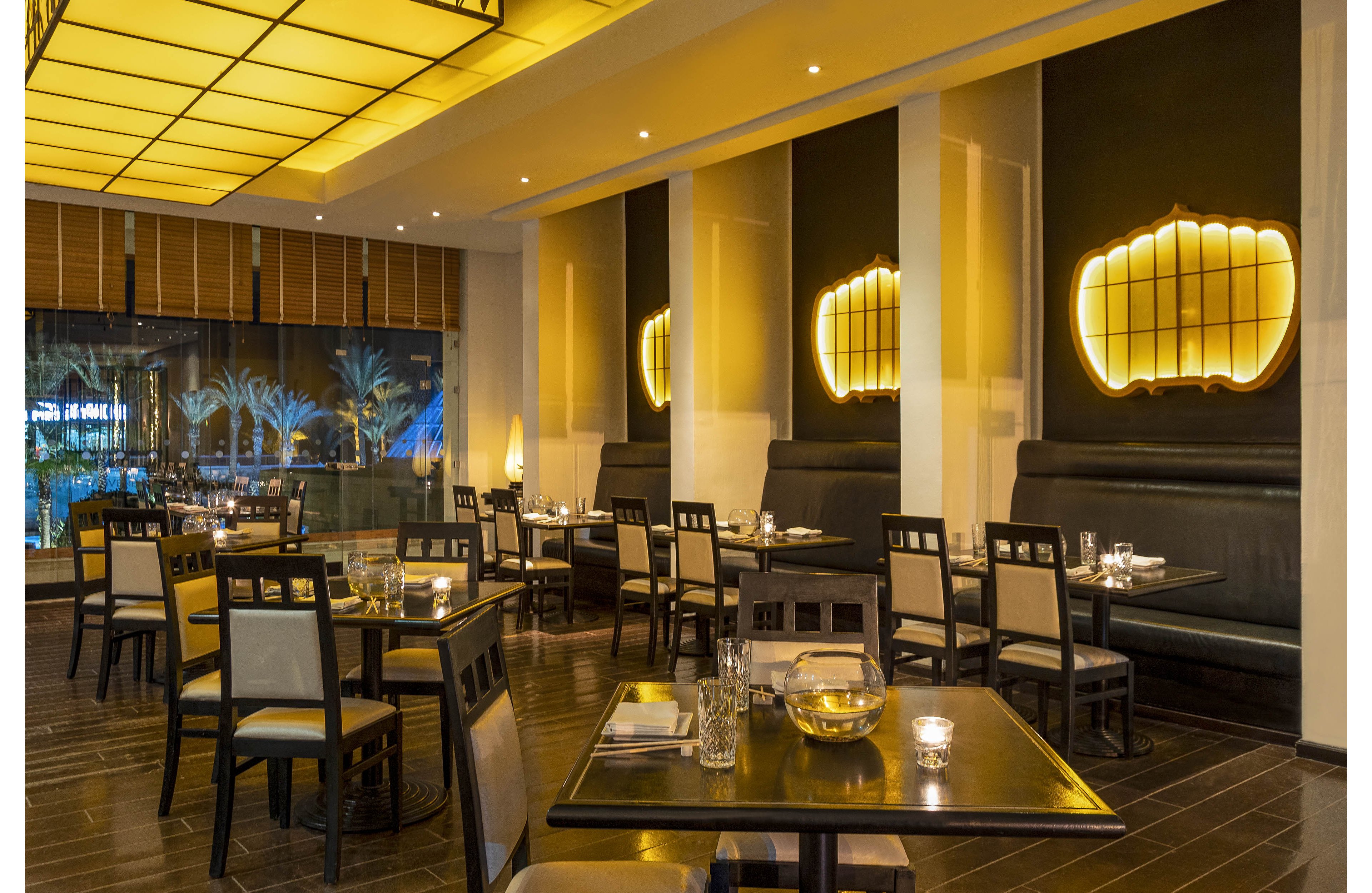 Contemporary restaurant dining area featuring neatly arranged tables and chairs. The setting is softly illuminated by large yellow ceiling panels and glowing wall fixtures. Glassware, candles, and simple table settings create an elegant yet relaxed atmosphere. Large windows reveal palm trees outside, adding a subtle tropical touch to the interior scene.
