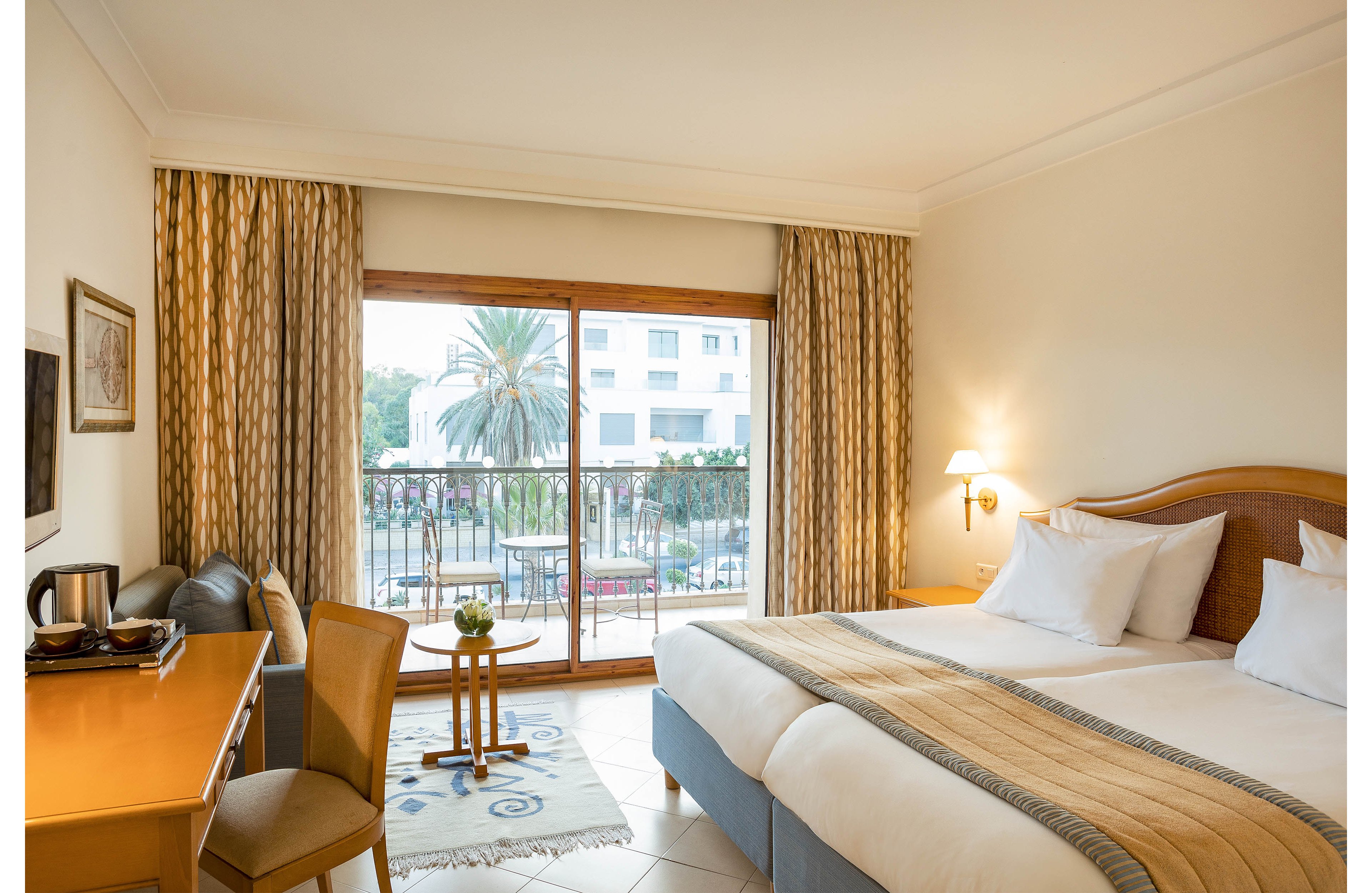 A neatly made hotel room features a large double bed with white linens and beige accents. A sliding glass door opens to a balcony overlooking palm trees and an outdoor pool area. A wooden desk with a chair, kettle, and cups sits opposite the bed, while patterned curtains frame the window. Warm wall lighting and neutral decor create a calm, inviting atmosphere for guests.