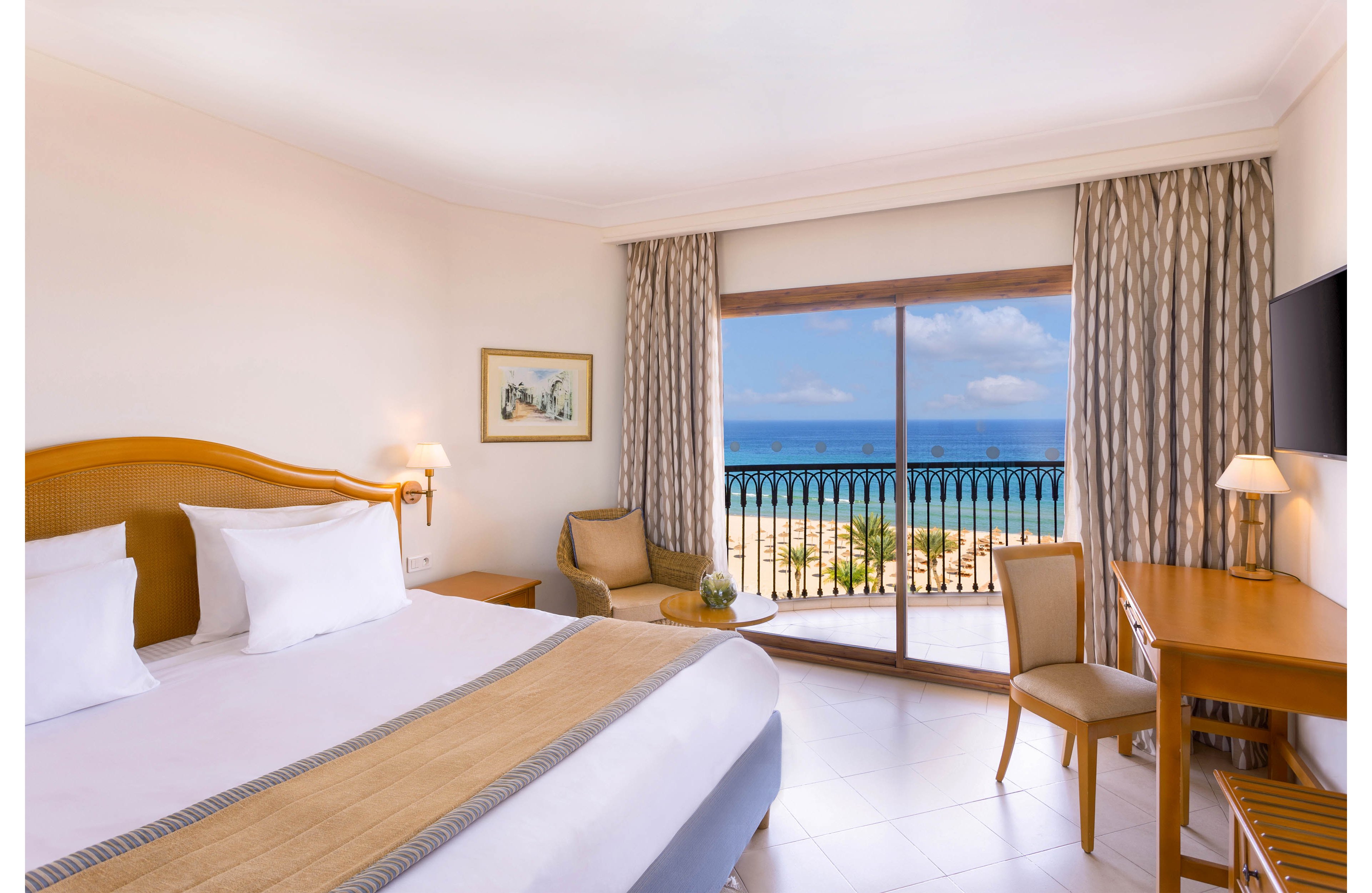 A spacious hotel bedroom features a large neatly made bed with white linens and neutral-toned accents. The room includes a wooden desk, chair, and small seating area beside the window. Floor-to-ceiling glass doors open to a balcony overlooking a sandy beach and blue ocean under a partly cloudy sky. Soft lighting and light-colored curtains create a calm, inviting atmosphere ideal for relaxation.