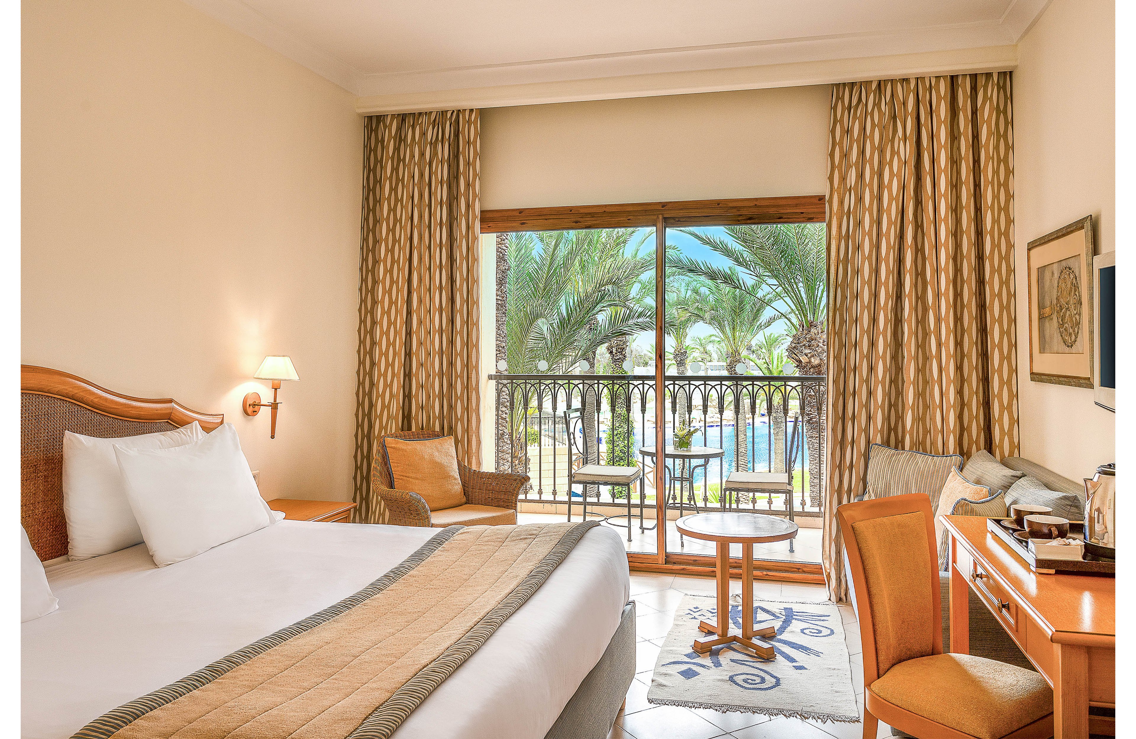 Bright hotel room with a large bed faces a balcony overlooking palm trees and a swimming pool. Warm wooden furniture, including chairs and a small round table, create a comfortable seating area near the window. A desk with a chair and refreshments sits to the right, adding functionality for work or leisure. Neutral walls and patterned curtains contribute to a calm, inviting resort atmosphere.