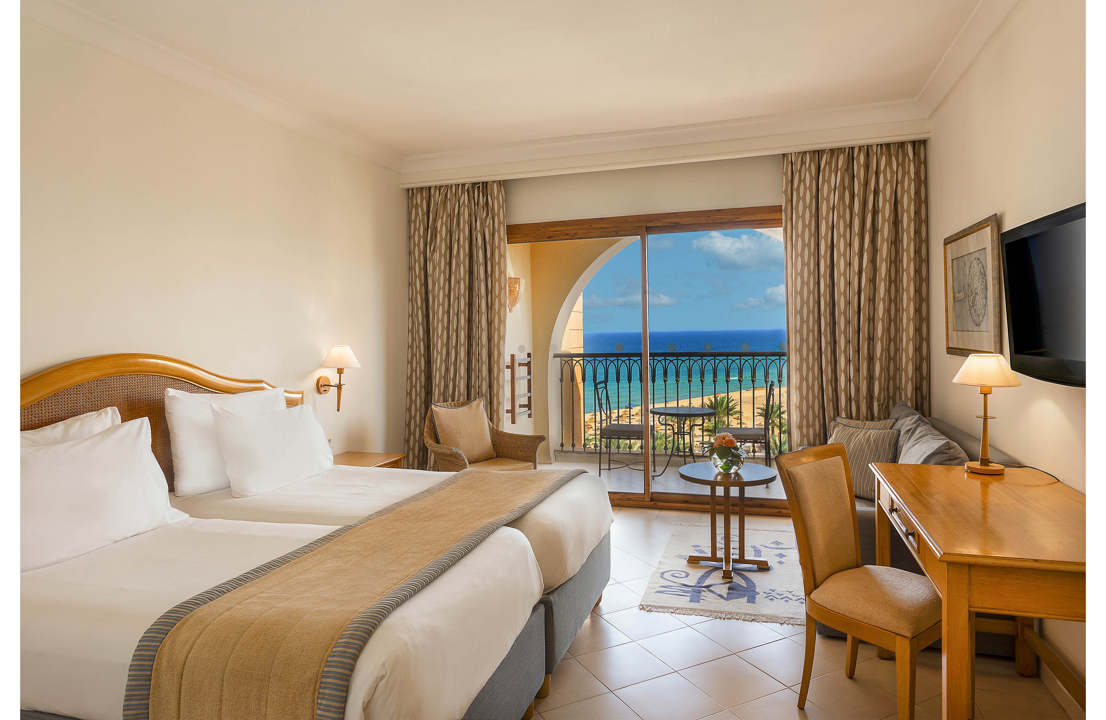 A neatly made hotel room features two twin beds with white linens and beige throws. The space includes a wooden desk, chair, and wall-mounted television on the right side. Large sliding glass doors open to a balcony with a table and chairs overlooking a blue ocean and clear sky. Neutral tones, soft lighting, and tiled floors create a calm, inviting atmosphere for guests.