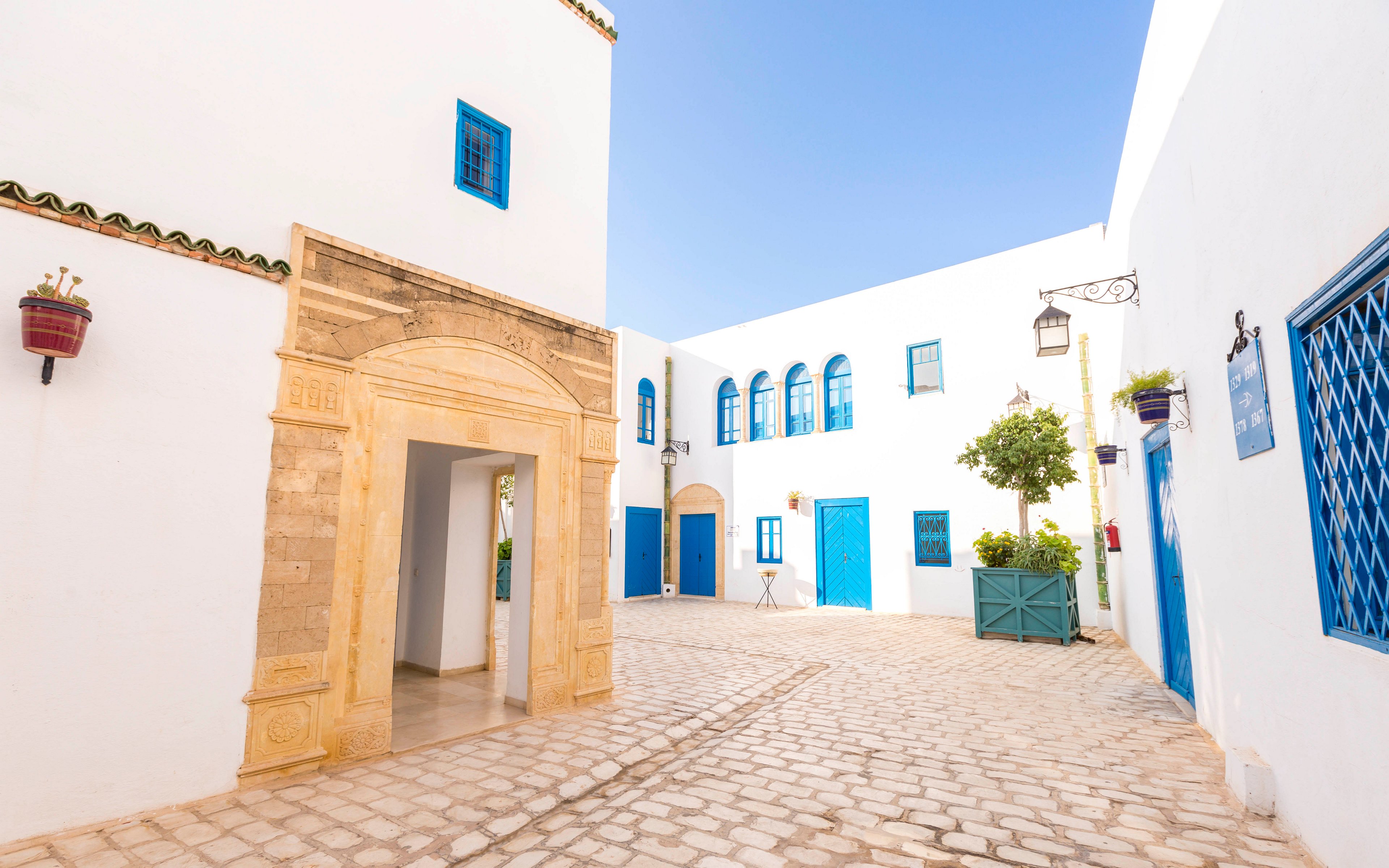 A bright white courtyard features traditional Mediterranean architecture with vivid blue doors and windows. The cobblestone ground leads past an ornate stone archway into the open space. Potted plants and small trees add greenery against the white walls. Warm sunlight and clear skies create a calm, inviting atmosphere in this quiet alleyway.