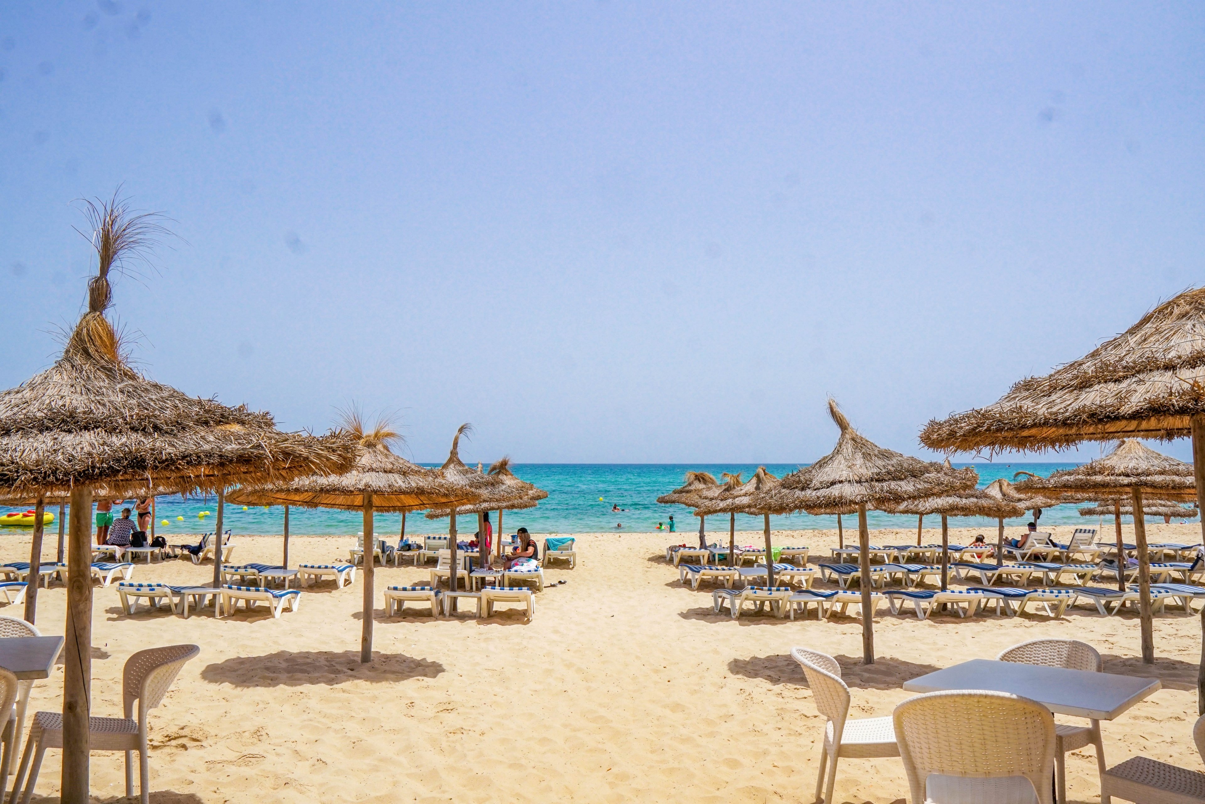A sandy beach is lined with rows of straw parasols and sun loungers facing the turquoise sea. White plastic chairs and tables sit in the foreground, suggesting a casual seaside café area. People relax under the umbrellas and near the shoreline, enjoying the bright, clear day. The calm water and cloudless sky create a tranquil, summery atmosphere.